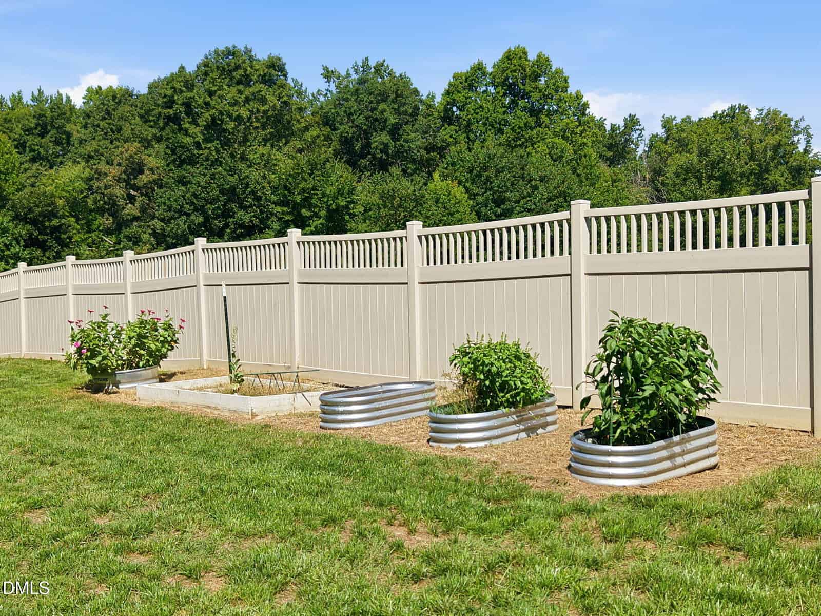 A backyard garden with three metal raised beds and some plants, next to a beige privacy fence. Green grass and trees are visible in the background under a blue sky.