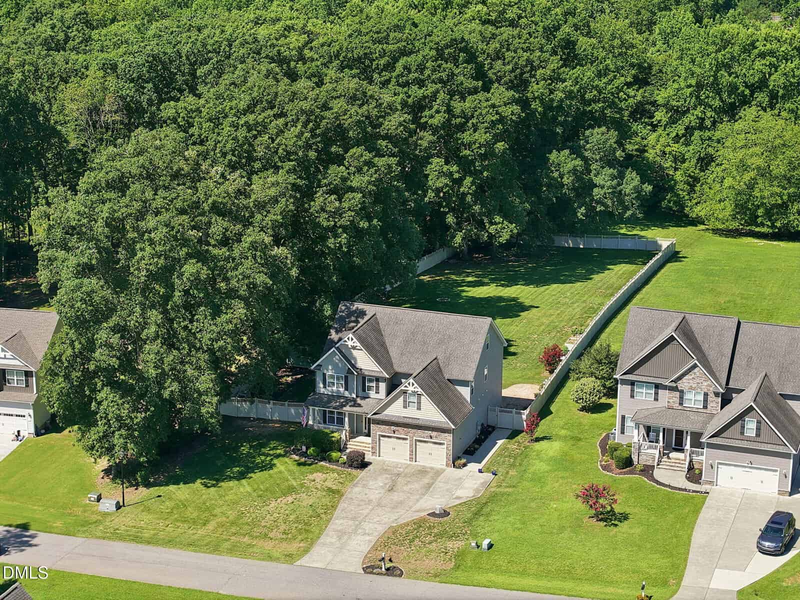 Aerial view of two suburban houses with driveways, front lawns, and a backyard surrounded by trees and a fence; a wooded area is visible behind the houses.