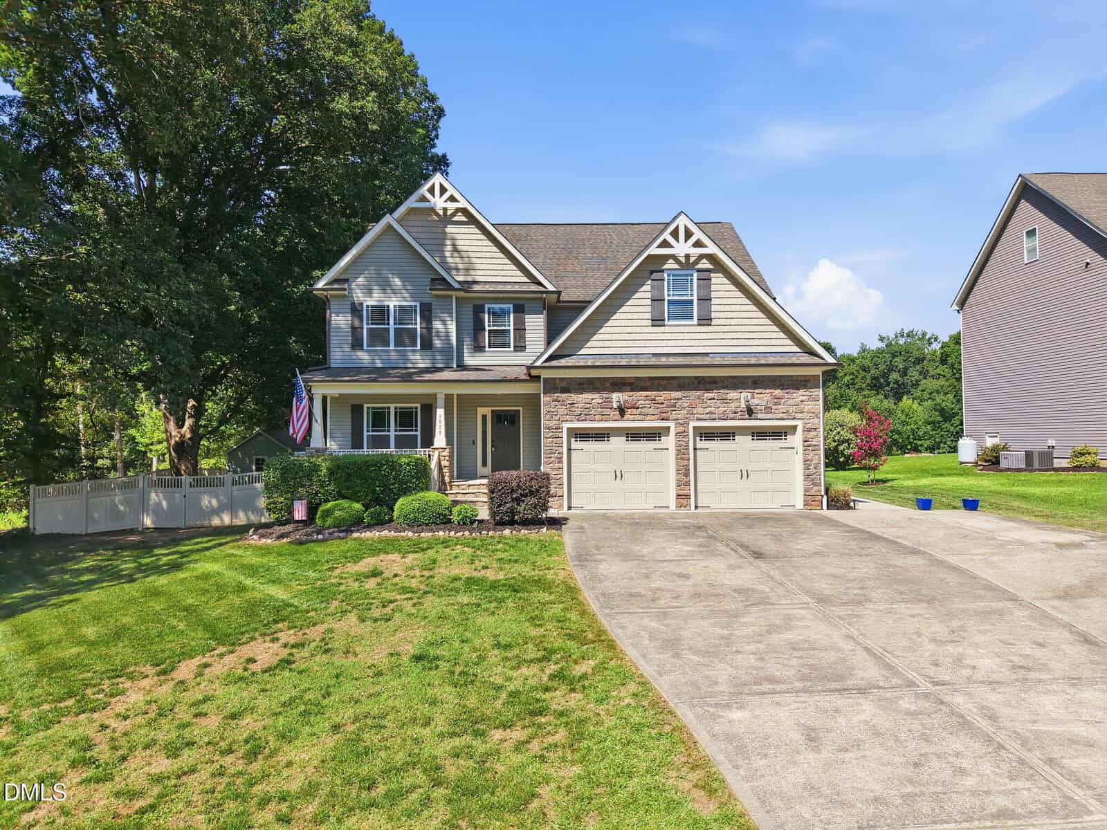 Two-story suburban house with beige siding, stone accents, and a double garage. American flag hangs by the front porch. Concrete driveway, landscaped yard, and a white fence on the left. Clear sky with trees and neighboring house nearby.
