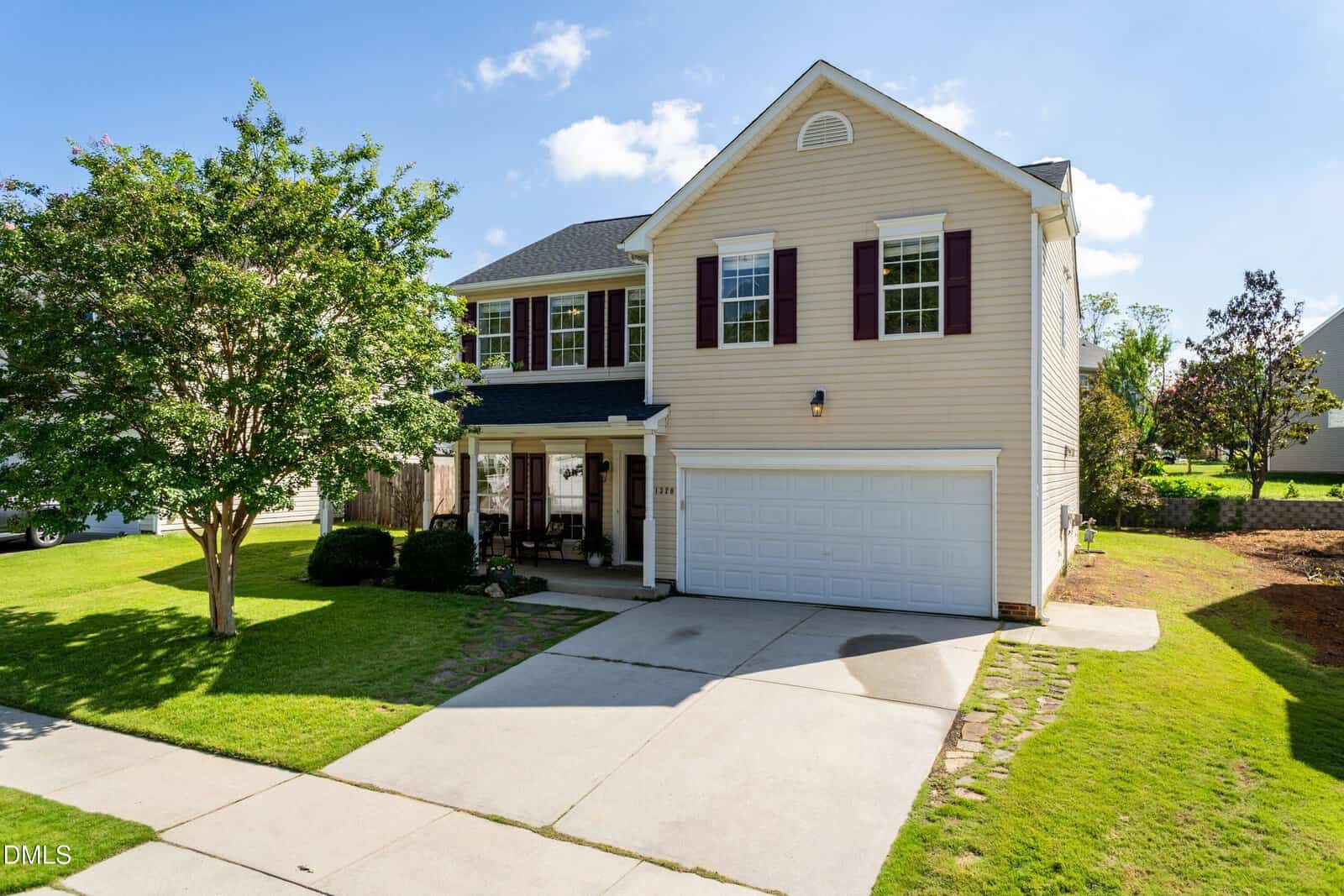Charming two-story beige house with white trim, dark shutters, and a double garage located at 1320 Marbank Street in Wake Forest. A covered porch with chairs sits to the left of the garage. The yard is grass with a few trees, and the driveway is concrete.
