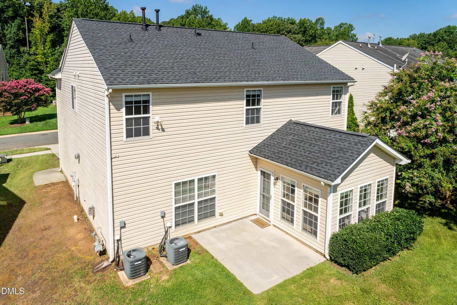 Beige two-story house with asphalt shingle roof, two HVAC units, a small concrete patio, and a sunroom extension. The property has minimal landscaping and is bordered by grass, trees, and a nearby street.
