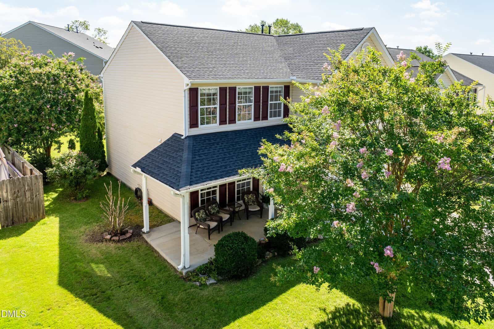 Two-story beige house with dark roof, red shutters, covered front porch, chairs, green lawn, and a flowering tree in the yard on a sunny day.