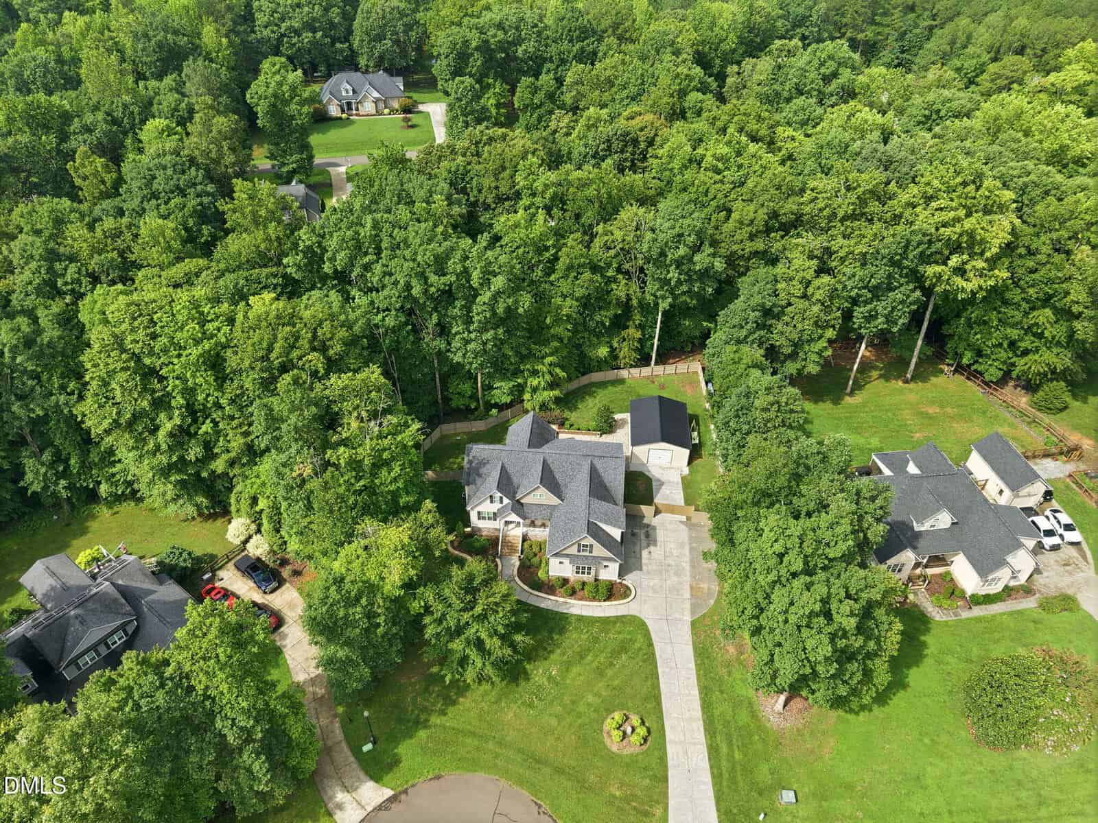 Aerial view of a large house with a circular driveway, surrounded by trees and greenery. Neighboring houses and a cul-de-sac are visible, with woods in the background. The scene appears to be a suburban neighborhood.
