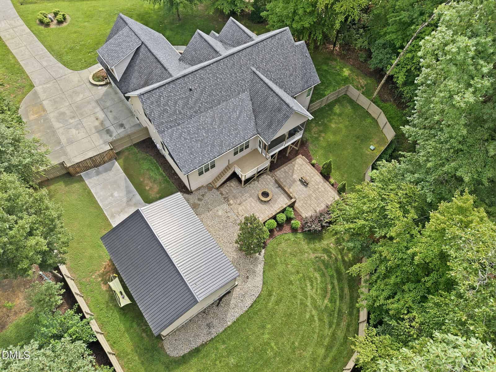 Aerial view of a large house with gray roof, detached garage, spacious driveway, fenced backyard, patio area with fire pit, and surrounding trees and lawn.
