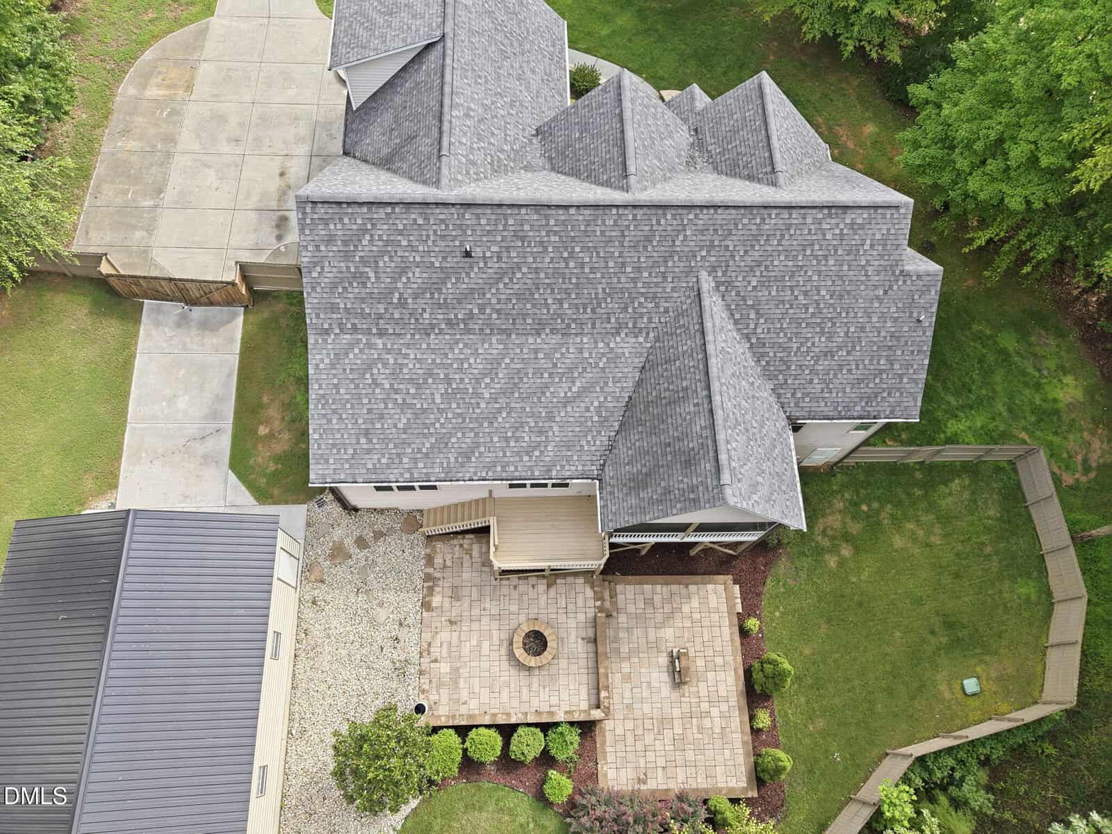 Aerial view of a house with a gray shingle roof, a wooden deck, a paved patio area with seating and firepit, a fenced backyard, green lawn, and adjacent driveway with a detached garage or shed.