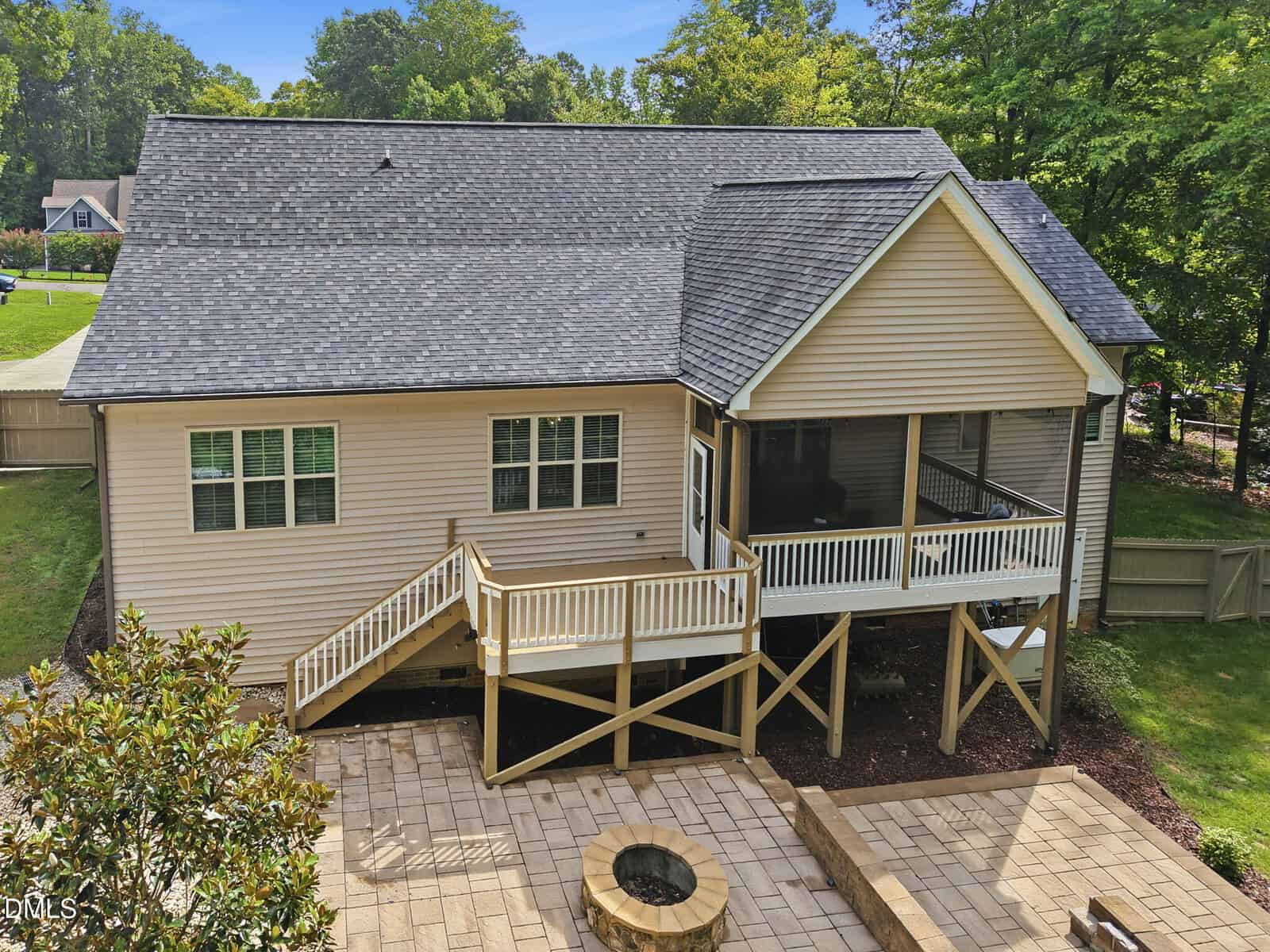 Back view of a beige house with a dark roof, raised wooden deck, screened porch, stairs, fenced yard, and stone patio with a fire pit. Green trees and a neighboring house are visible in the background.