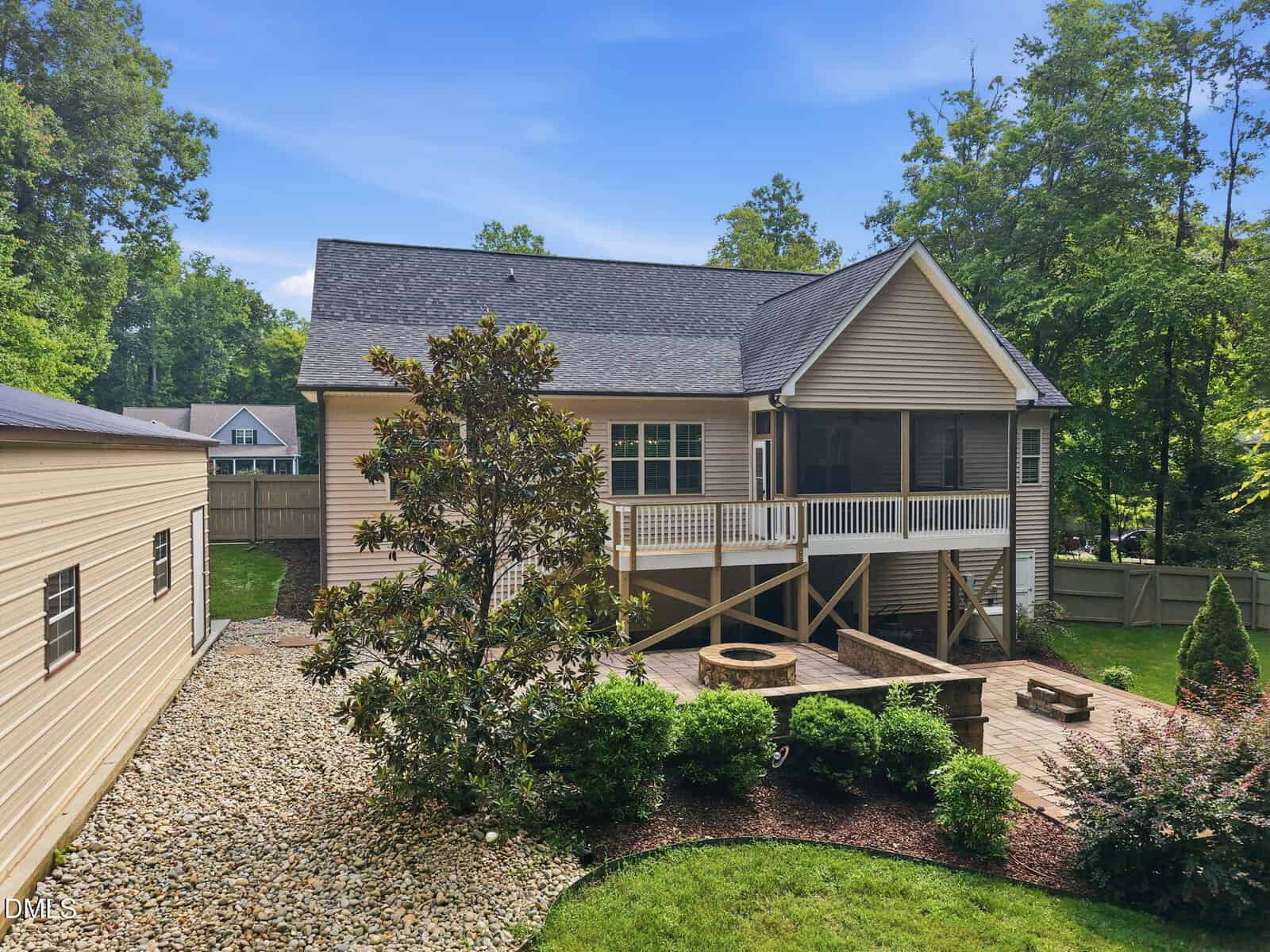 Backyard with a beige house, elevated screened porch, stone patio with a firepit, landscaping, small shrubs, a gravel area, and a detached garage, surrounded by a wooden fence and trees under a blue sky.