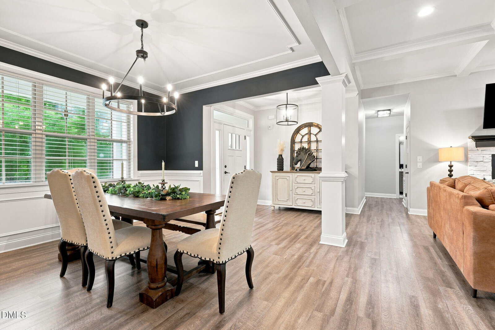 Modern dining area with a wooden table, beige upholstered chairs, greenery centerpiece, large window with blinds, and chandelier. Off 1096 Buggy Lane, the hallway leads to a beige sofa and lighted wall decor in an open, bright home interior.