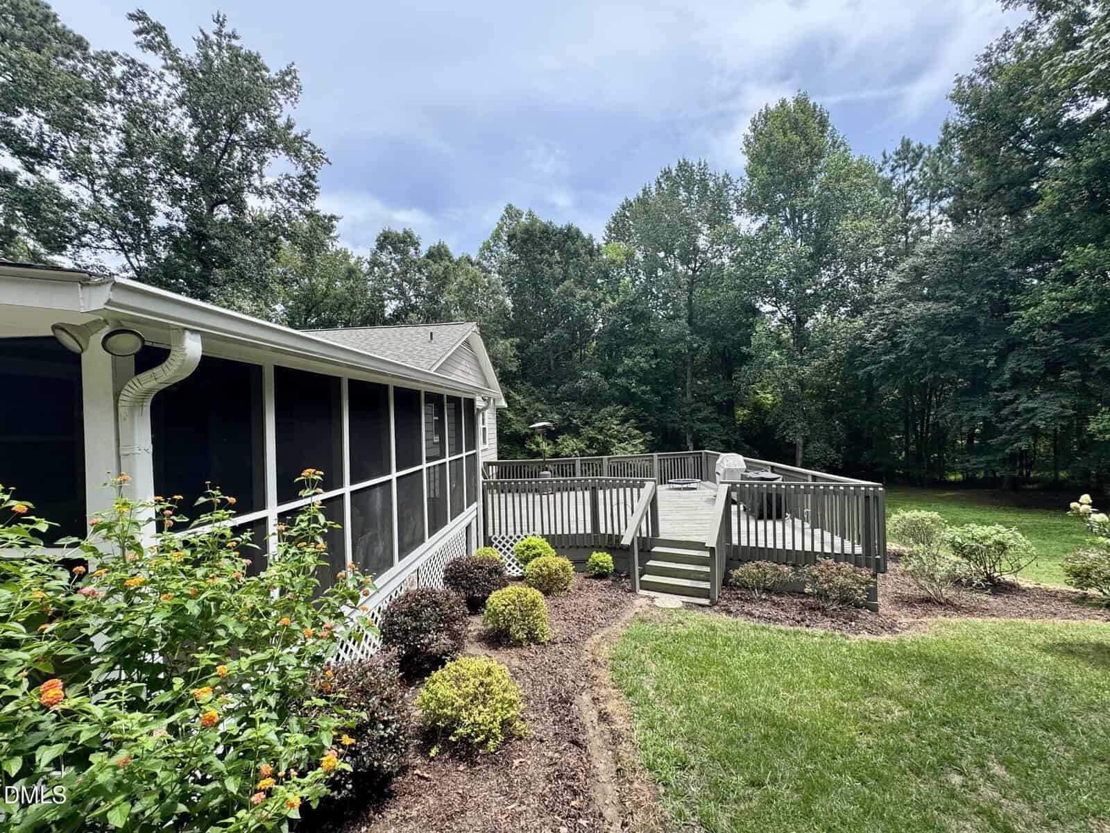 A house with a screened porch and a raised wooden deck, featuring stairs leading to a grassy backyard bordered by trees and landscaped bushes under a partly cloudy sky.