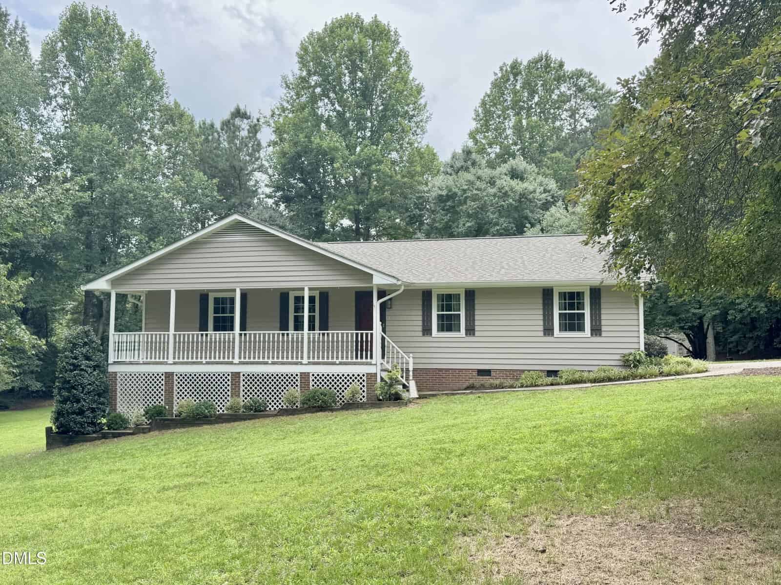 Single-story house with light gray siding, white trim, and a covered front porch located at 1384 Faulkner Town Road in Henderson. The home sits on a grassy yard with shrubs and is surrounded by tall trees. The sky is partly cloudy.
