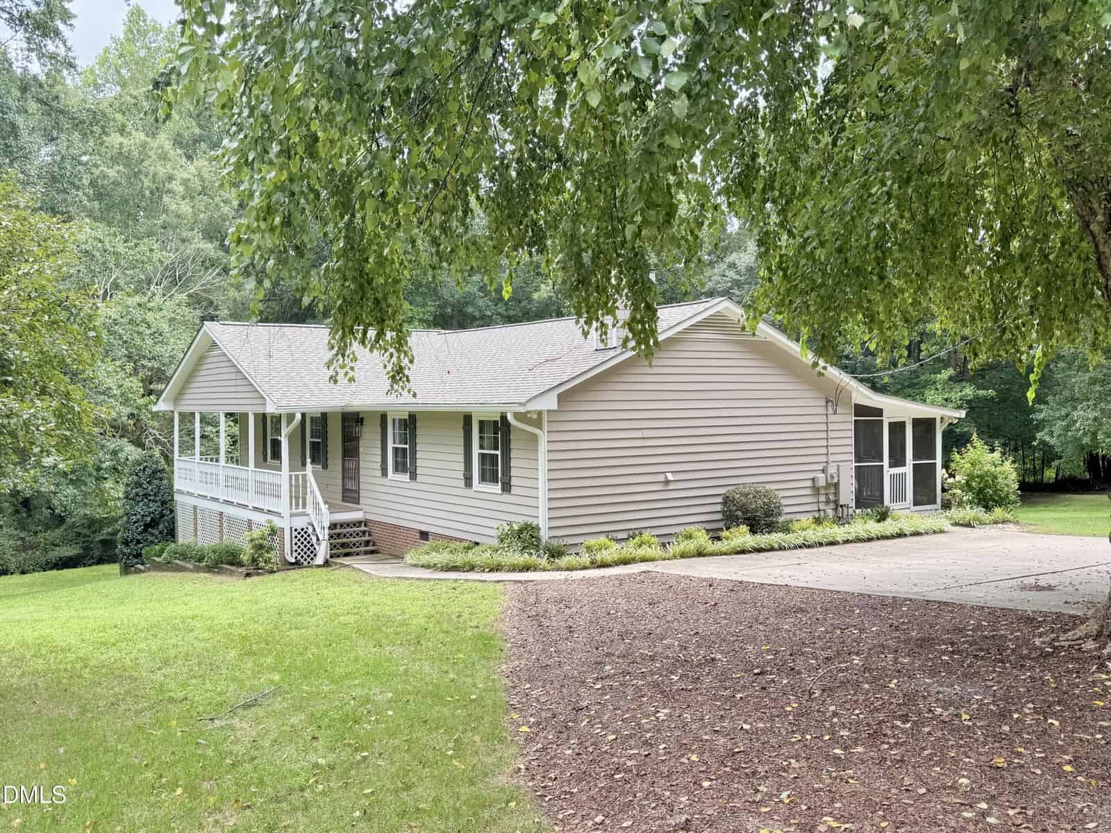 Single-story beige house with a front porch, white railings, and screened side porch, surrounded by trees and grass. A concrete driveway leads to the side entrance. The setting appears suburban or semi-rural.