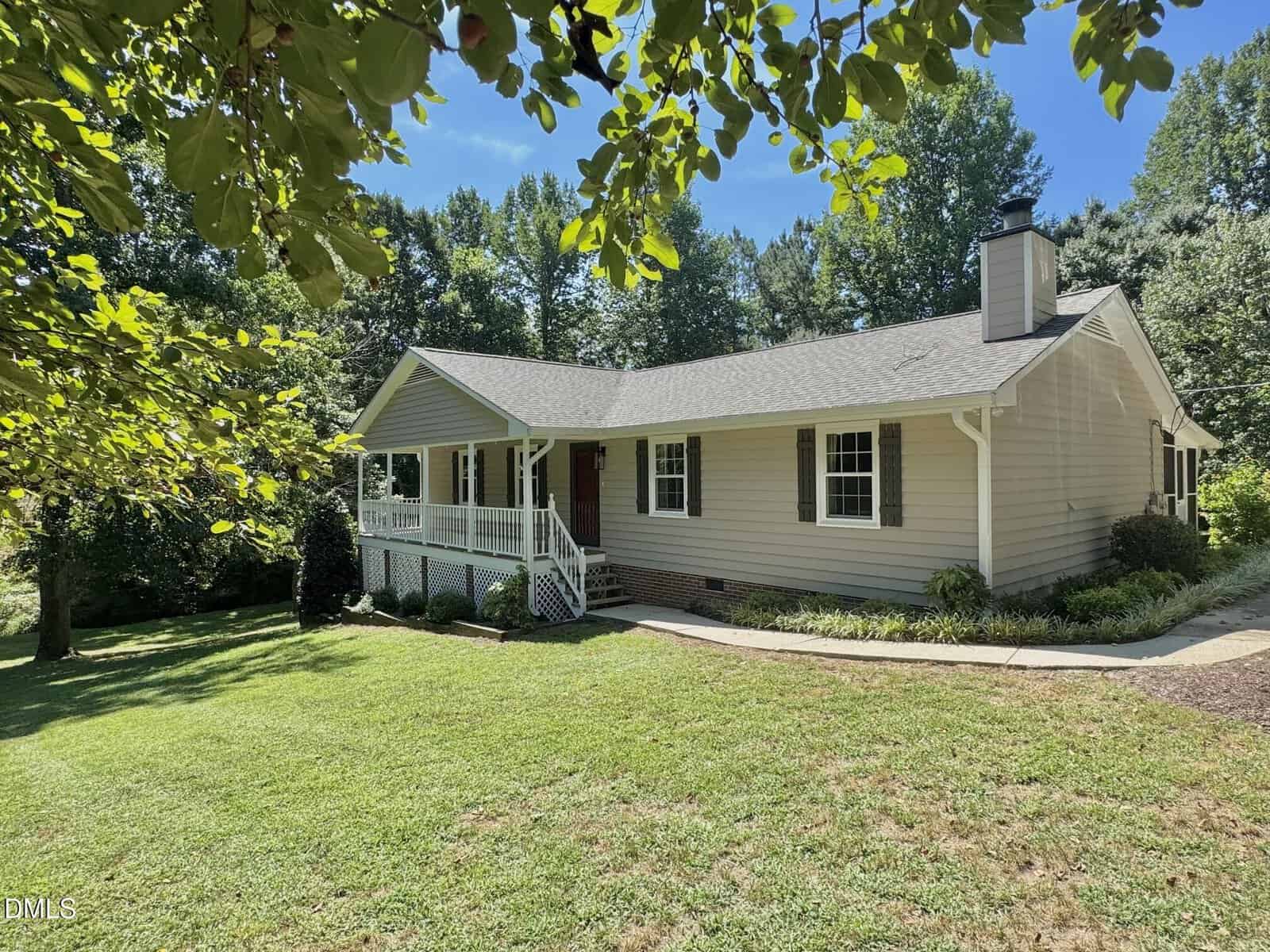 Single-story beige house with a front porch, white railing, and a brick foundation, surrounded by a grassy yard and trees under a sunny blue sky.