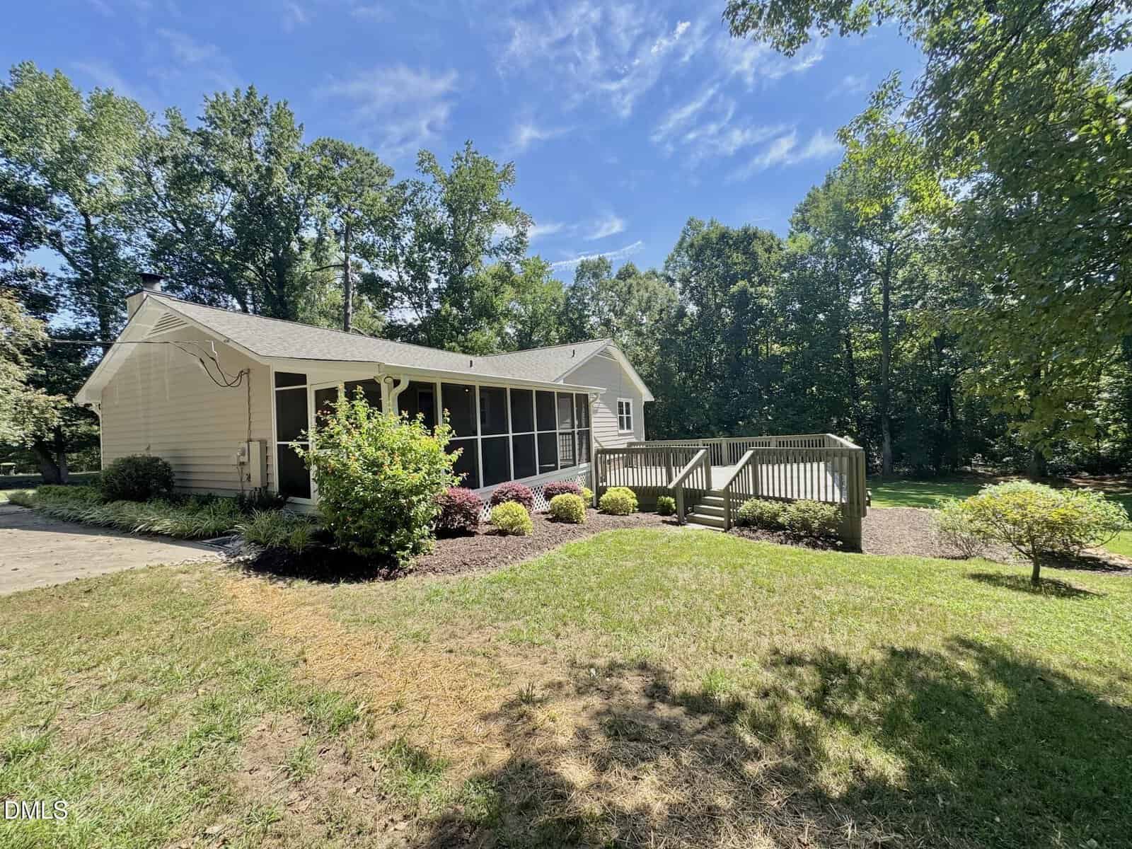 Single-story beige house with a screened porch, wooden ramp, and landscaped yard. Surrounded by green trees under a sunny blue sky.