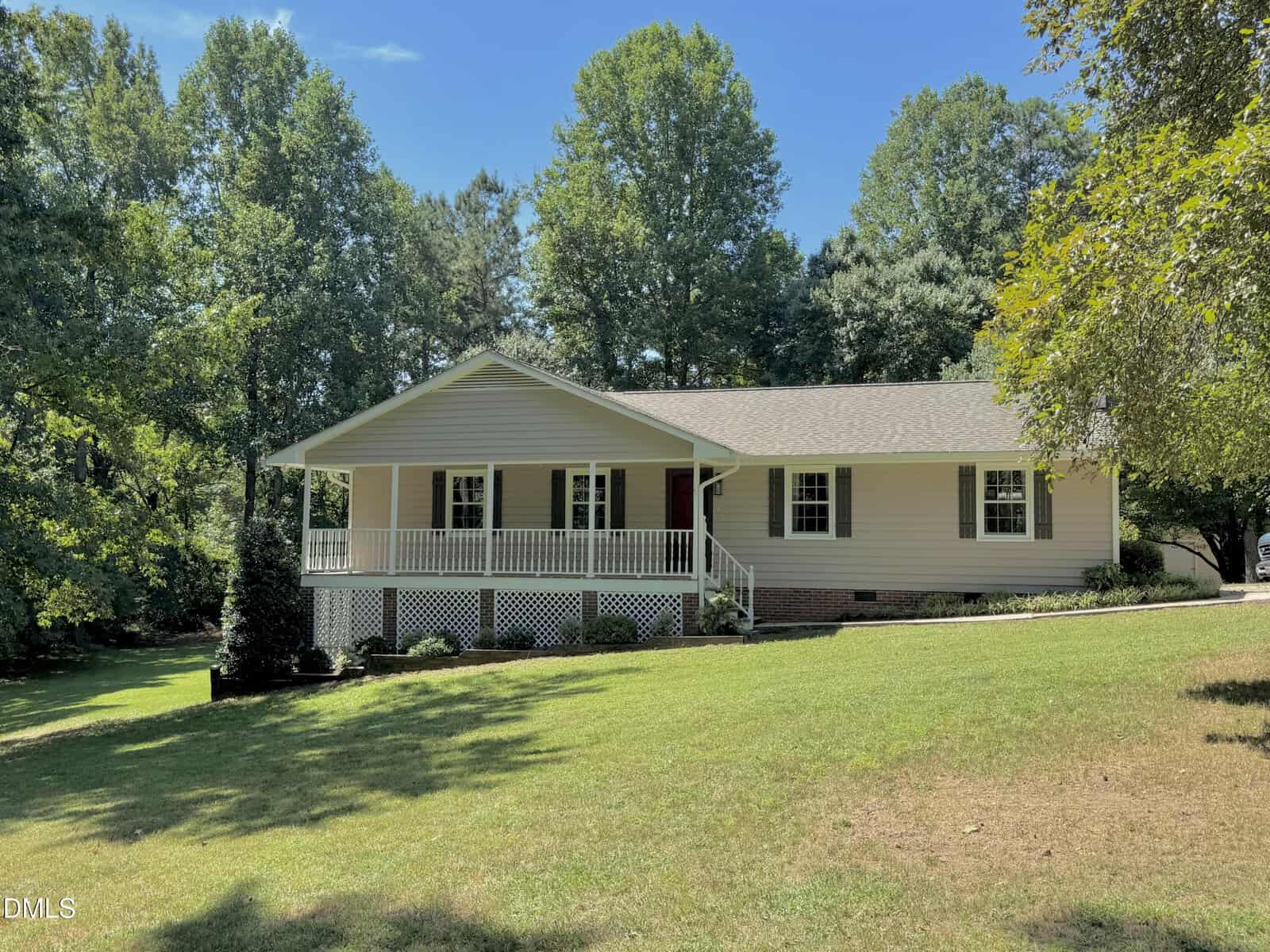 Single-story beige house with a large front porch, white railings, and lattice skirting, surrounded by green grass, mature trees, and a bright blue sky—nestled on 1384 Faulkner Town Road in Henderson.