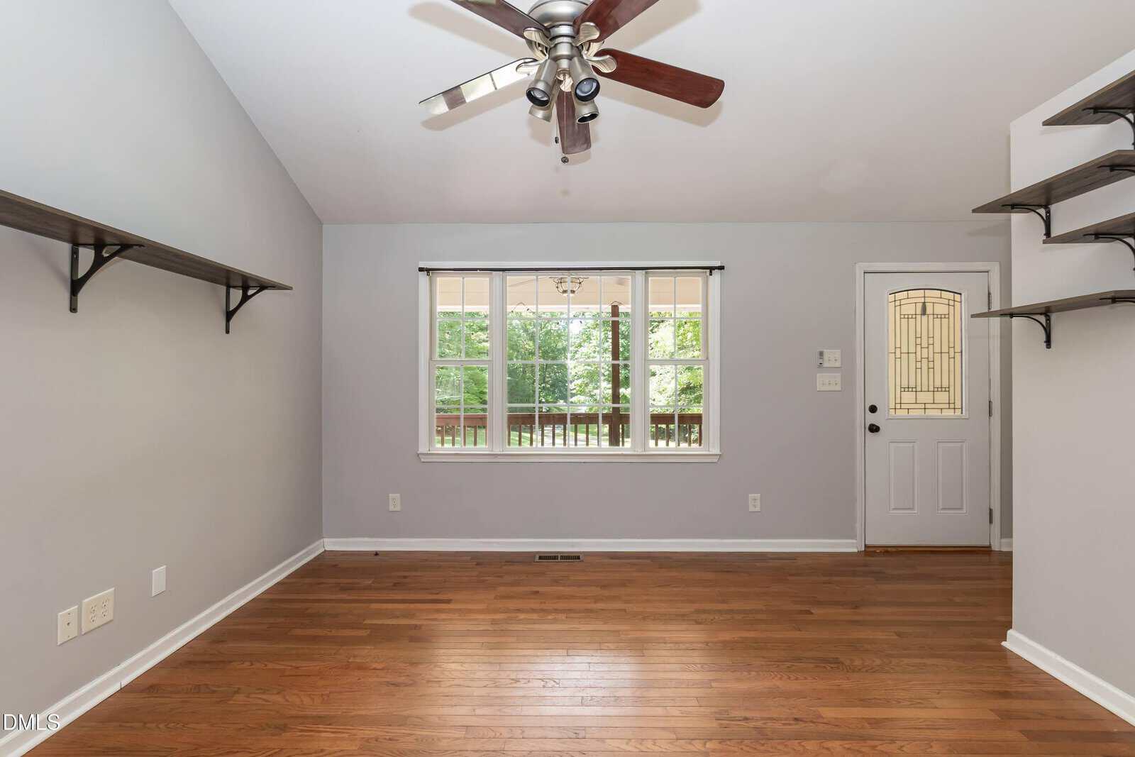 Empty room with light gray walls, hardwood floor, two wall-mounted shelves, ceiling fan with lights, large window, and a white door with a decorative glass panel. Trees are visible outside the window.