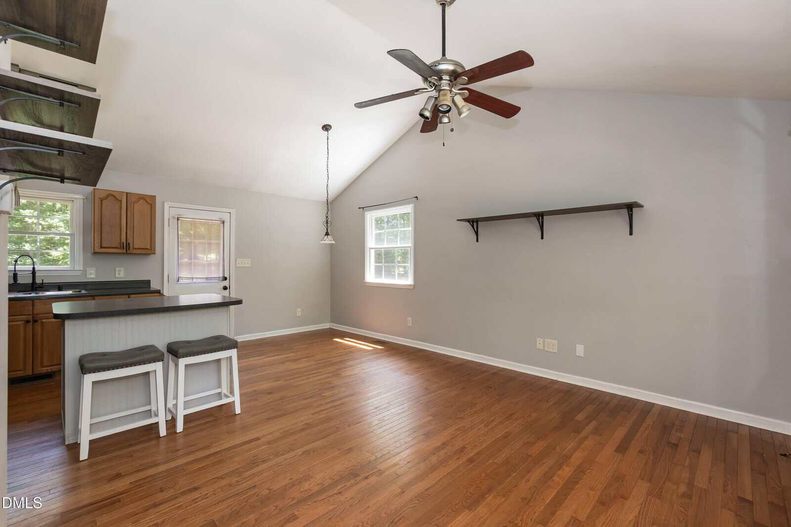 A bright, empty room with hardwood floors, a ceiling fan, a kitchen counter with two stools, open shelves, and a wall-mounted shelf under a window. Light gray walls and natural light from windows.