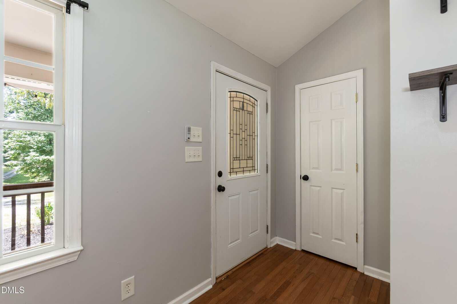 A small entryway with a white front door, a matching closet door, light gray walls, wood flooring, and a window showing trees outside.