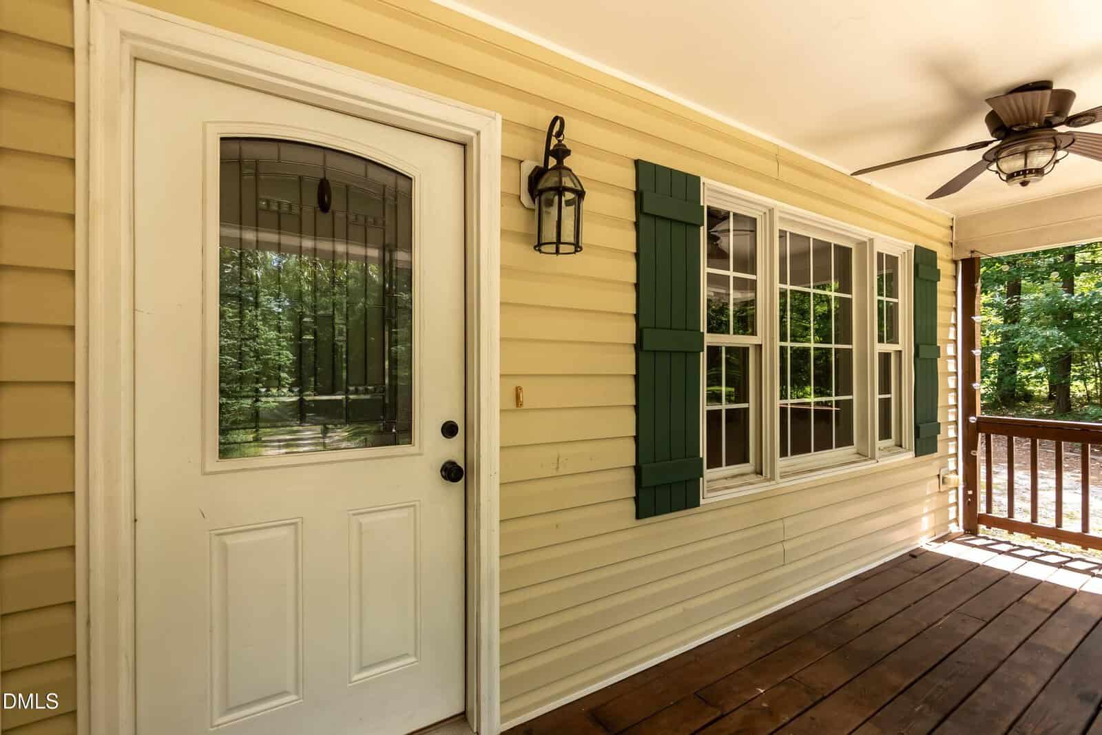 A covered porch with a white door, a lantern-style light fixture, three double-hung windows with green shutters, beige siding, a ceiling fan, and wooden flooring. Trees are visible through the windows and railing.
