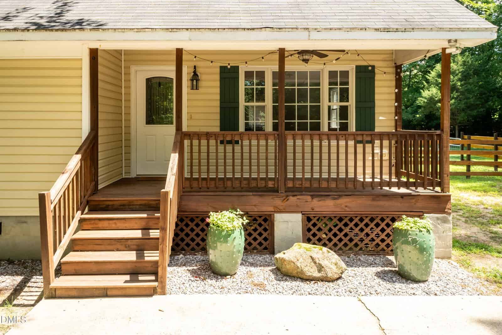 A front porch with wooden steps and railing, light yellow siding, a white door, and two green shutters flanking a window. Two large green planters and a rock sit by the steps. Trees and a wooden fence are in the background.