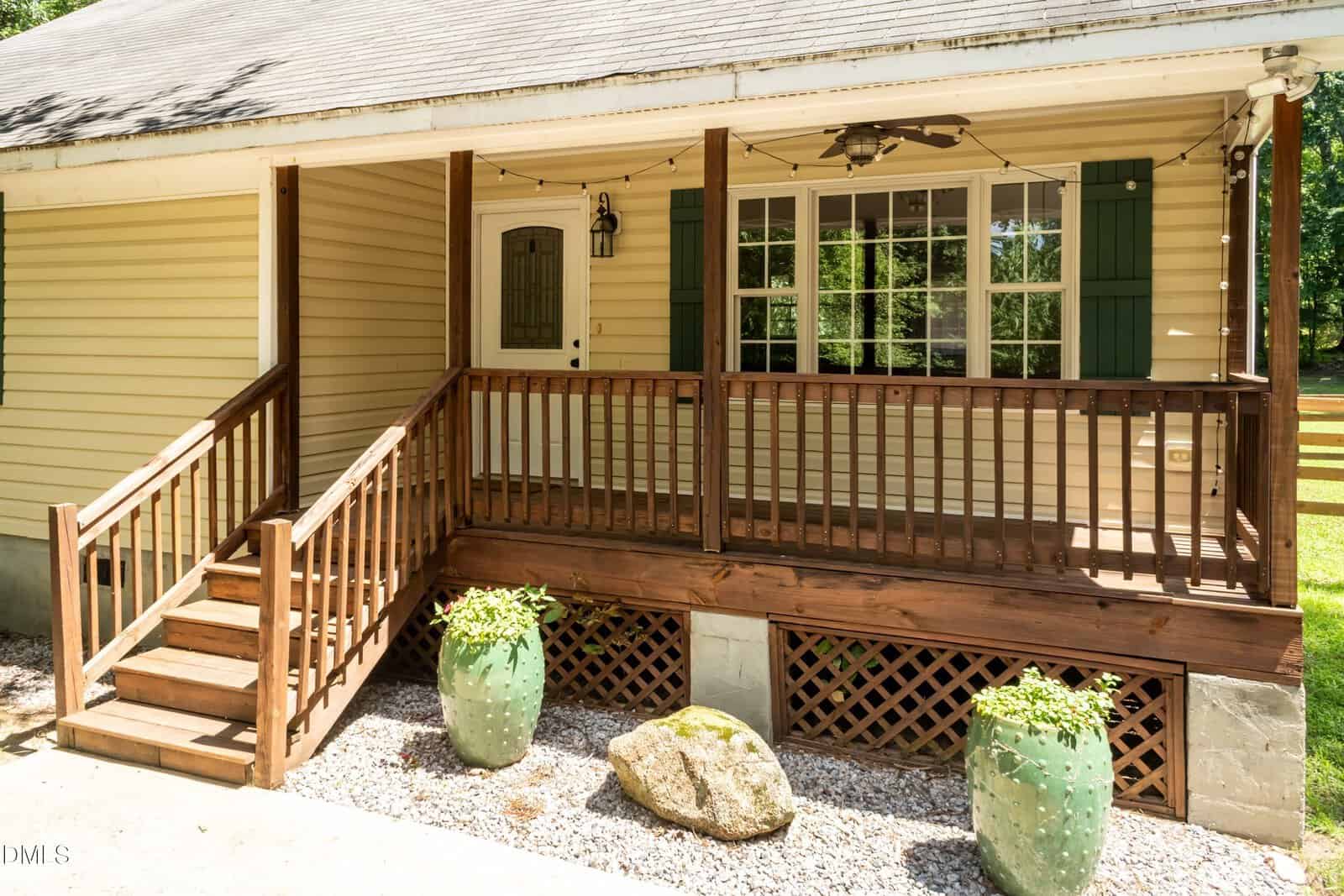 A wooden porch with steps and railing, featuring string lights, a ceiling fan, green shutters, large windows, two potted plants, and a large rock in front. The siding is yellow, and the porch area is partially shaded.