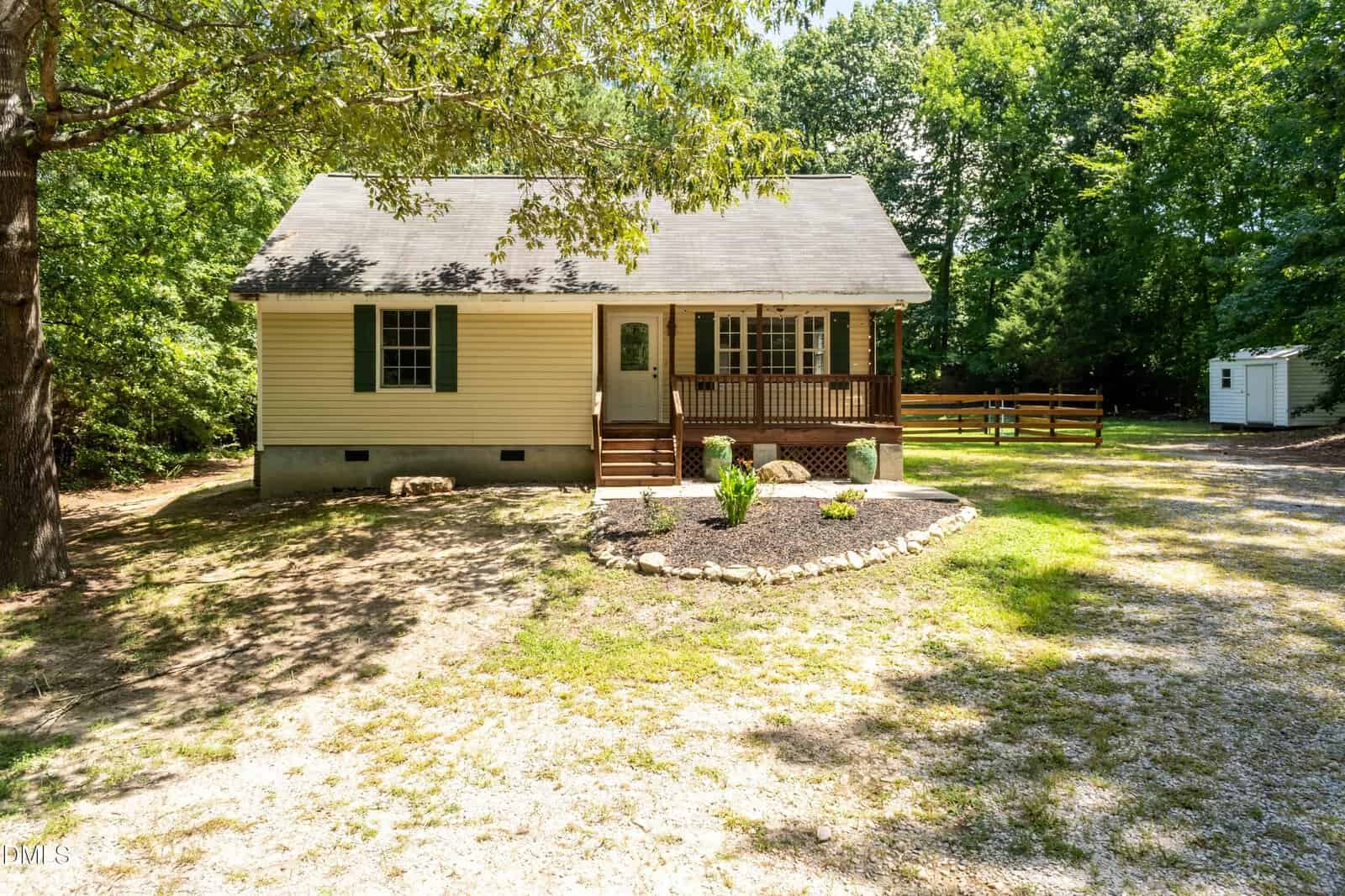 A small, single-story house with beige siding and a front porch sits among trees. There is a gravel driveway, a mulched garden bed with plants, and a fenced area in the background. Sunlight filters through the leaves.