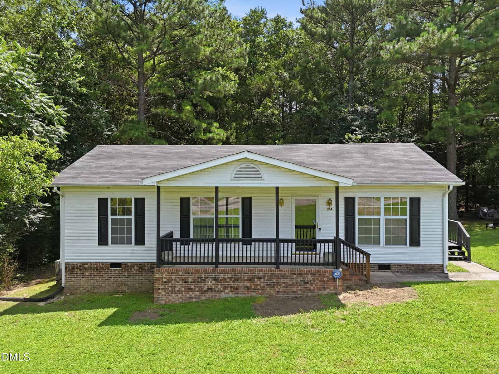 A single-story white house with black shutters, a brick foundation, and a covered front porch stands at 104 Pine Forest Drive in Henderson, surrounded by green grass and trees under a blue sky.