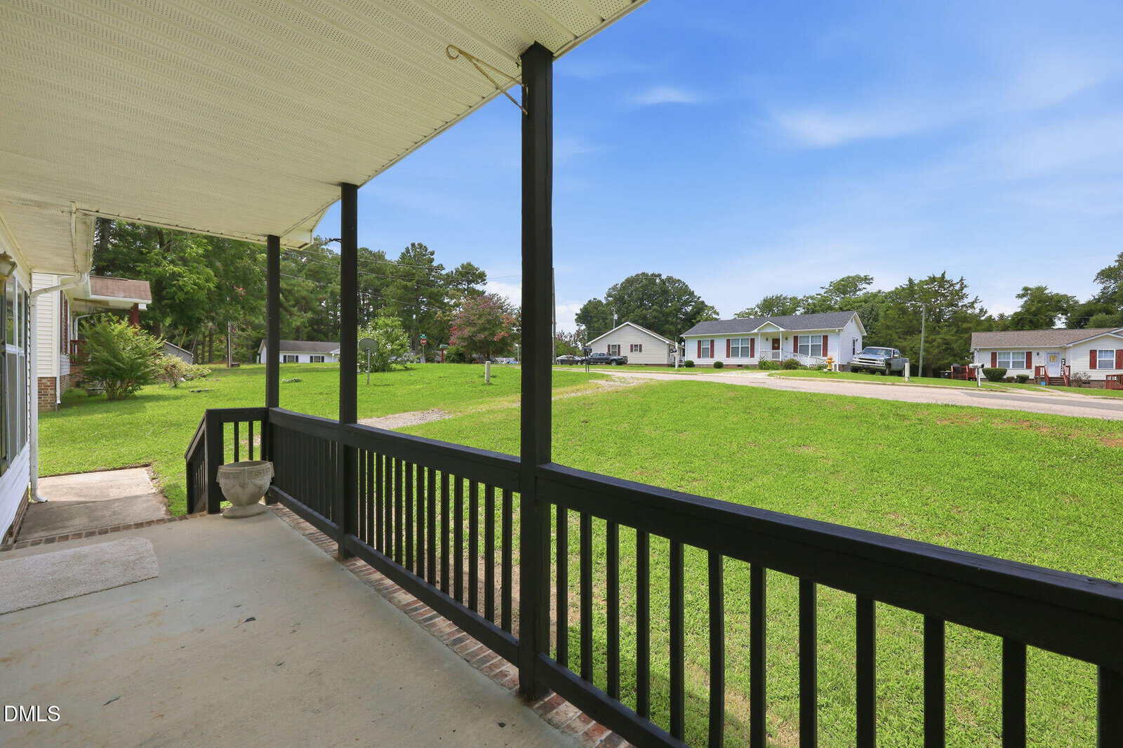 View from a covered porch with black railing, looking out onto a grassy yard and a suburban neighborhood with single-story houses under a clear blue sky.