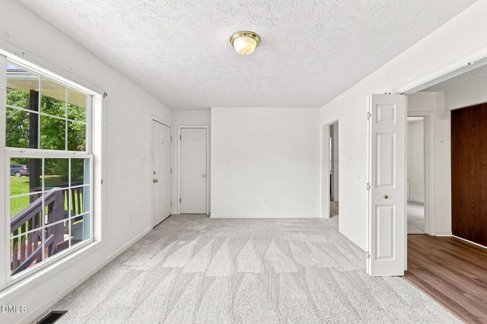 Bright, empty room with white walls, fresh light carpet, large window showing green yard, ceiling light, and double doors opened to an adjacent room with wood flooring.