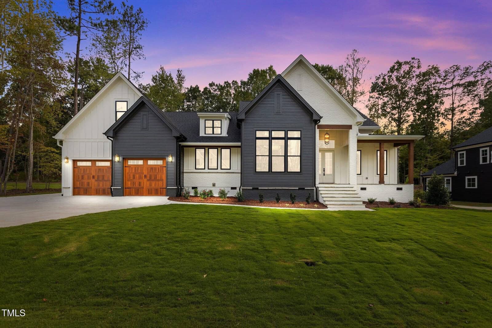 Modern two-story house at 106 Red Cardinal Court, Youngsville, featuring a white and dark gray exterior, large windows, covered front porch, and two wooden garage doors, surrounded by a green lawn and trees at sunset.