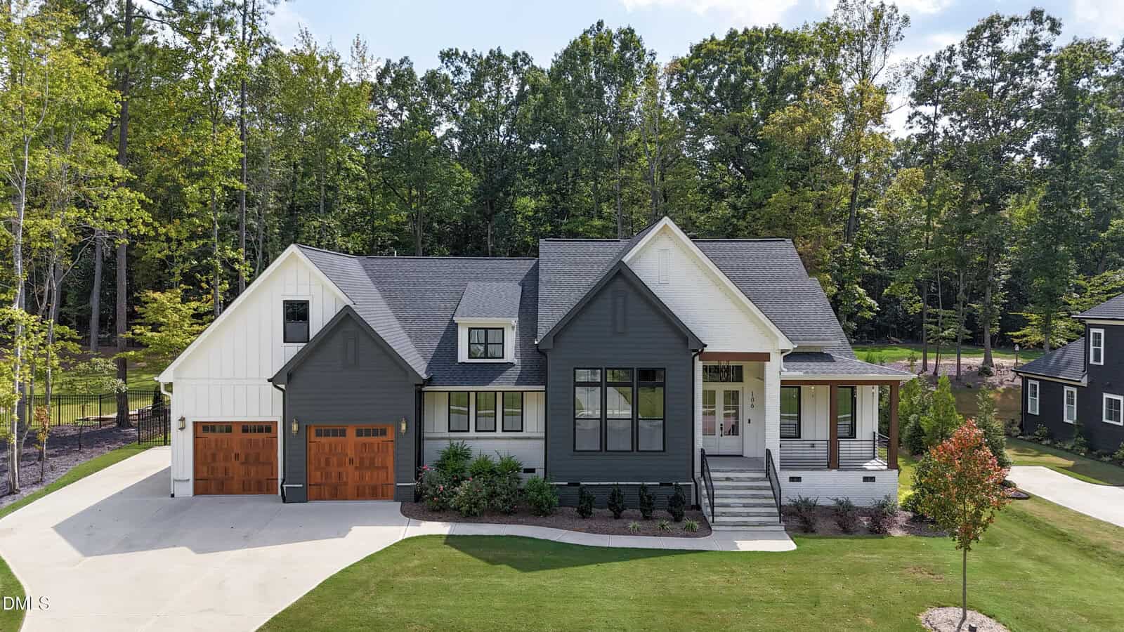 A modern two-story house with gray and white siding, large front windows, a covered porch, and two wooden garage doors, set among trees with a well-maintained lawn and concrete driveway.