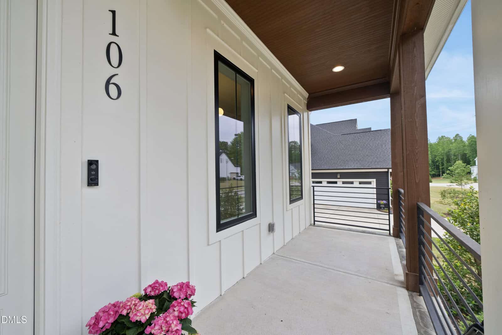 A modern house porch with white vertical paneling, large black-trimmed windows, house number 106, a black railing, pink flowers in a pot, and a view of a driveway and detached garage in the background.