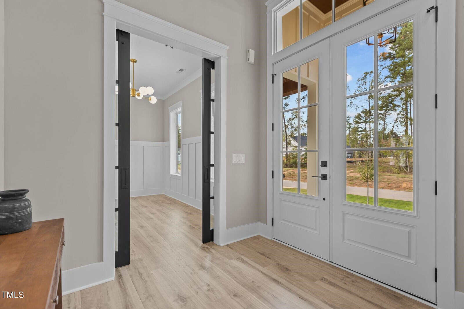 A bright entryway with double glass front doors, light wood flooring, neutral walls, and an open view into a room with a black-trimmed opening, white wainscoting, and a modern chandelier.