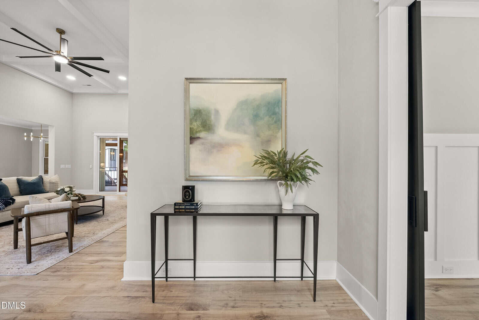 A narrow black console table with a plant, books, and decor sits against a light wall under a landscape painting. The space has wood flooring and leads to a living area with sofas and a ceiling fan.