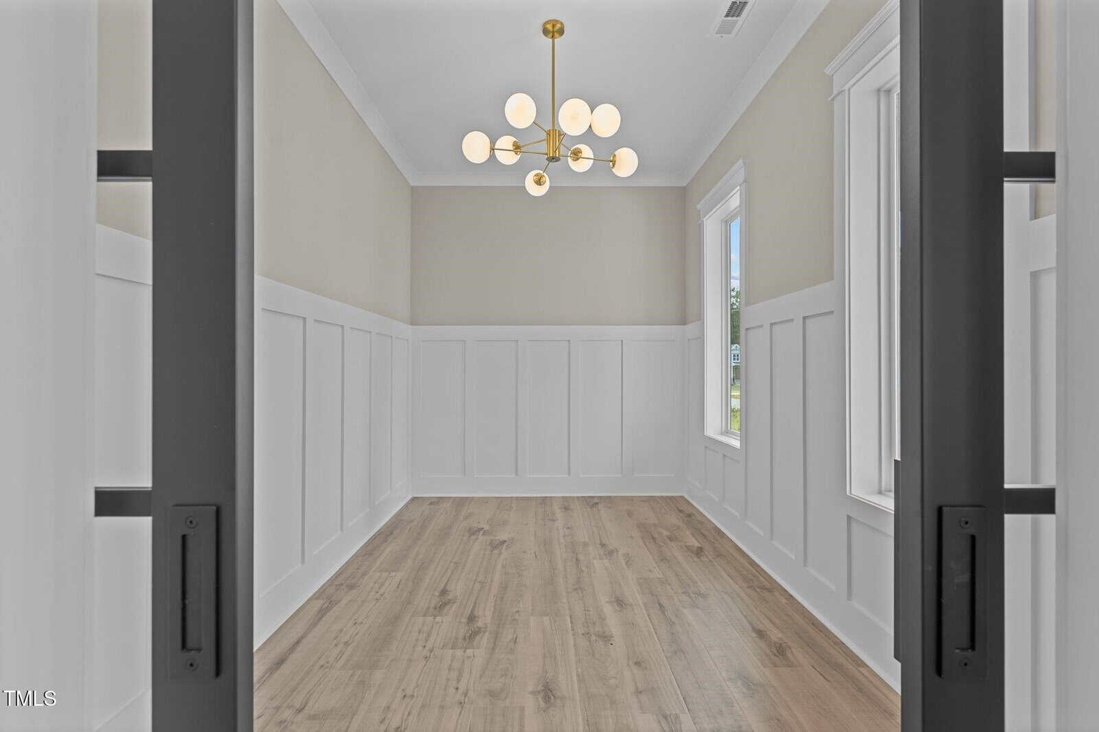 A bright, empty room with light wood flooring, white wainscoting walls, tall windows, and a modern gold chandelier hanging from a high ceiling.