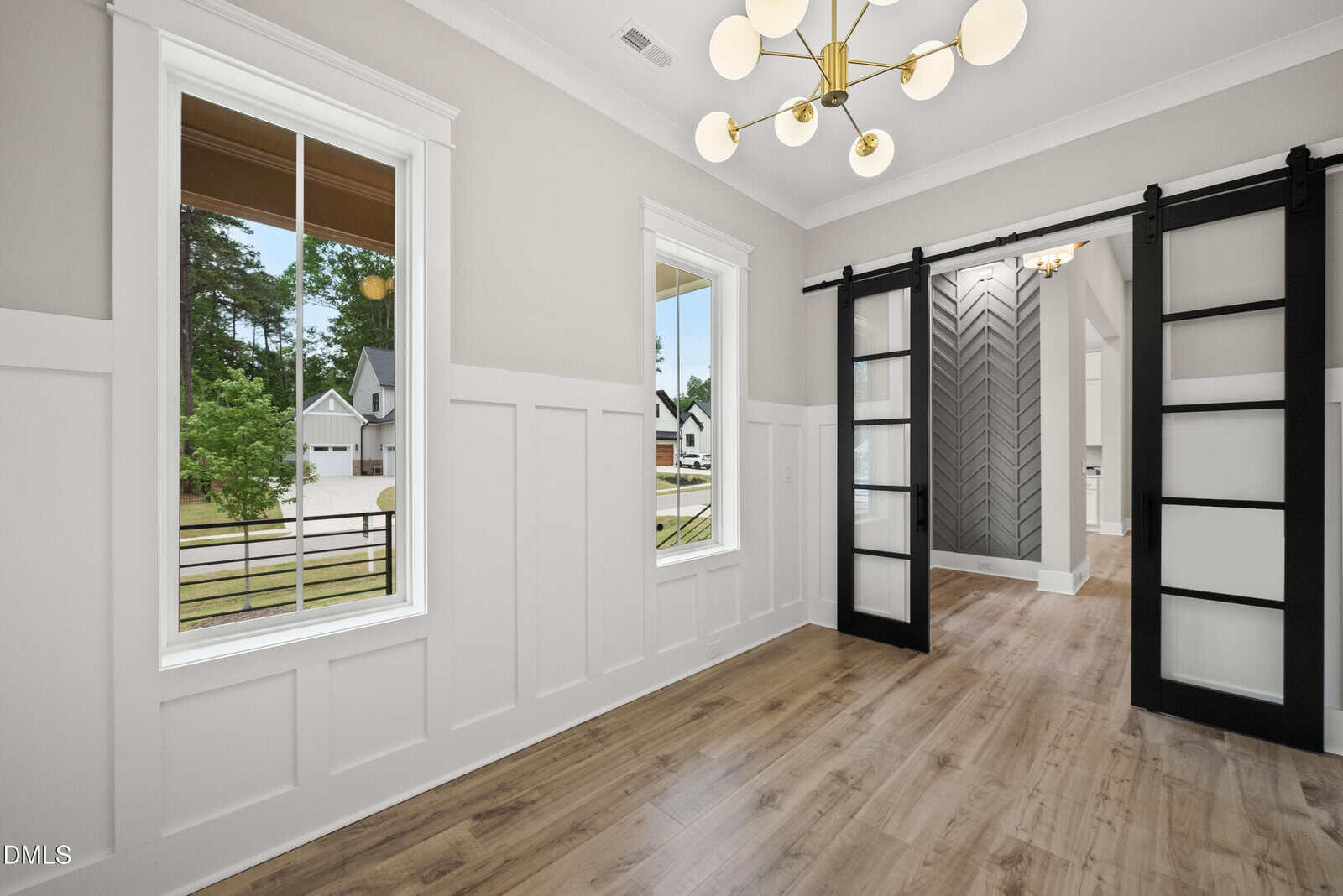 Bright room with large windows, light wood floors, white wainscoting, and a modern chandelier. Black sliding barn doors open to a hallway with chevron-patterned accent wall. Houses and trees are visible outside.
