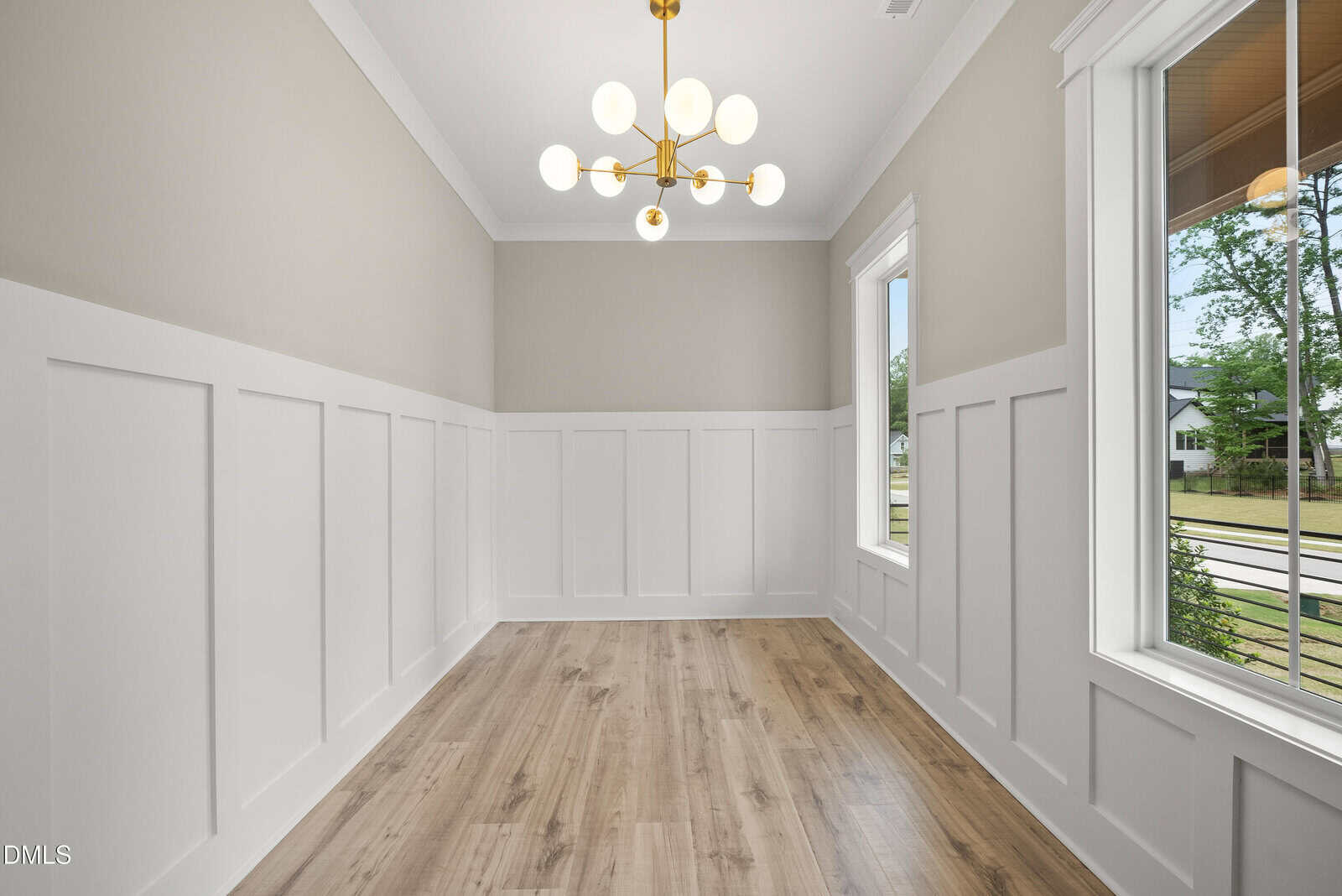 A bright, empty room with light wood floors, white wainscoting, beige upper walls, large windows, and a modern chandelier hanging from a white ceiling.