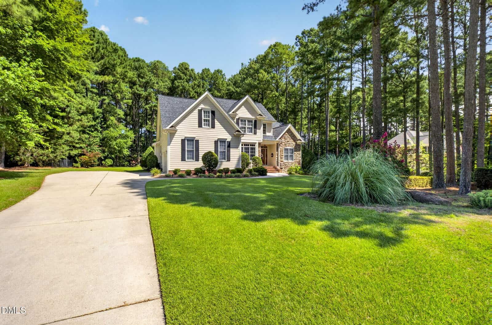 A beige two-story house with dormer windows sits surrounded by tall trees. A wide driveway leads to the home, and the front yard is landscaped with green grass and shrubs under a clear blue sky.