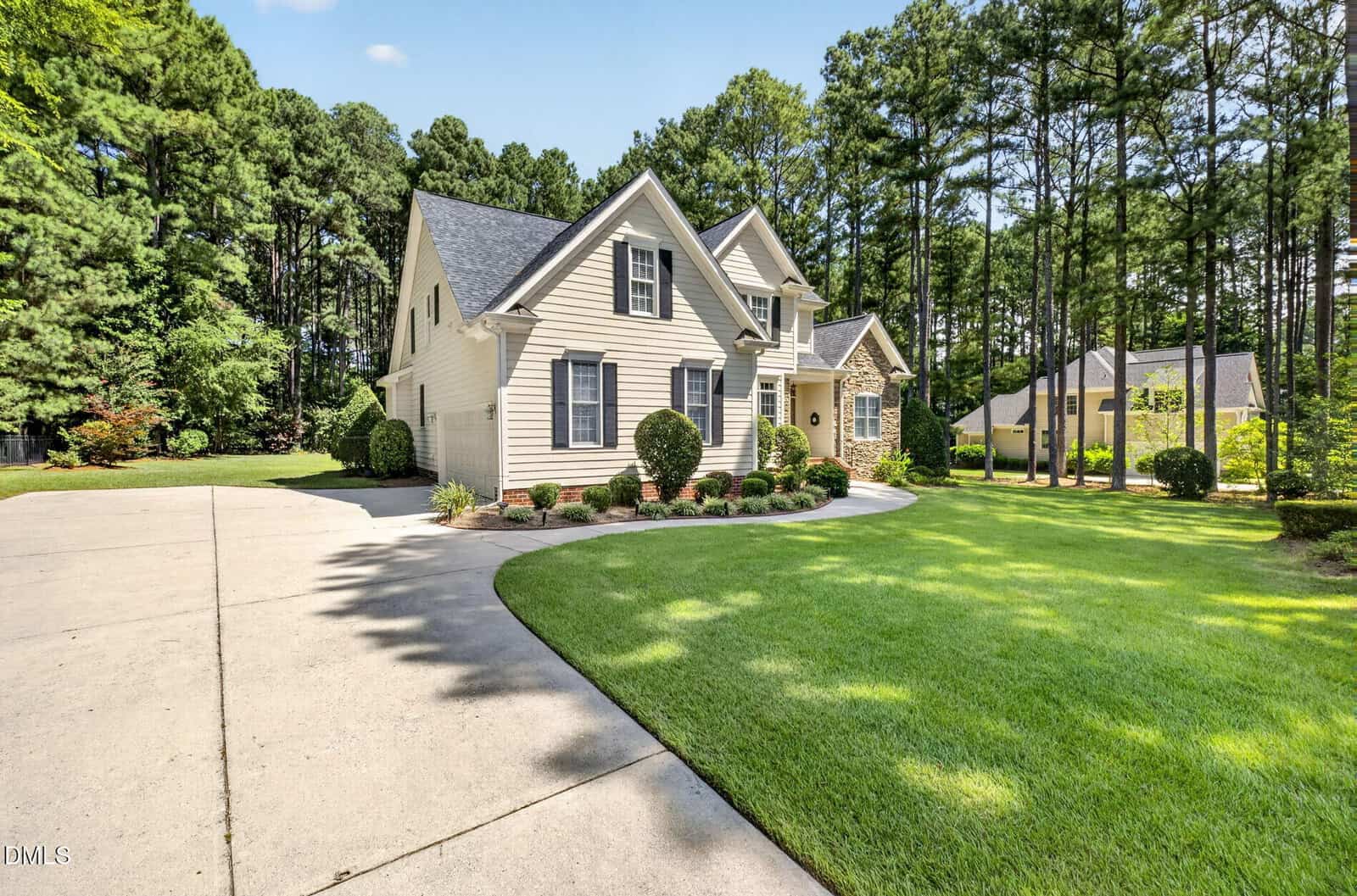 A two-story beige house with stone accents, gabled roof, and dormer windows sits next to a neatly trimmed lawn and a curved driveway, bordered by tall trees and landscaped bushes.