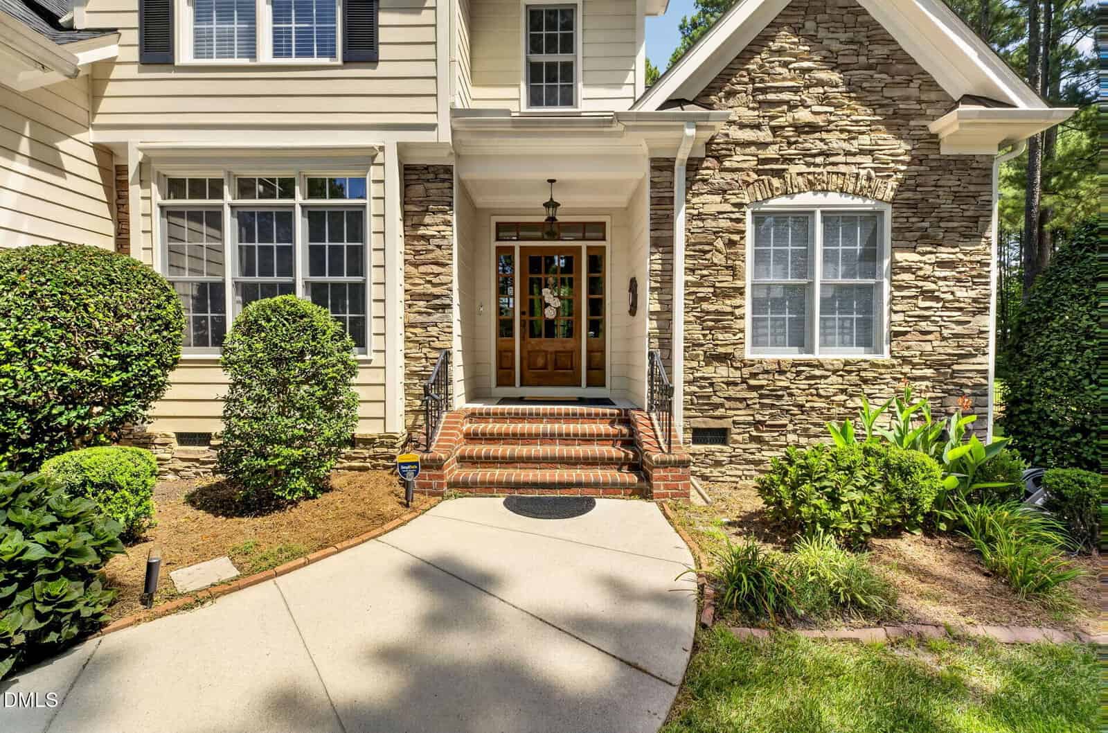 Front entrance of a house with beige siding, stone accents, white trim, and a wooden door with glass panels. Brick steps lead to the door. Manicured bushes and plants border a curved concrete walkway.