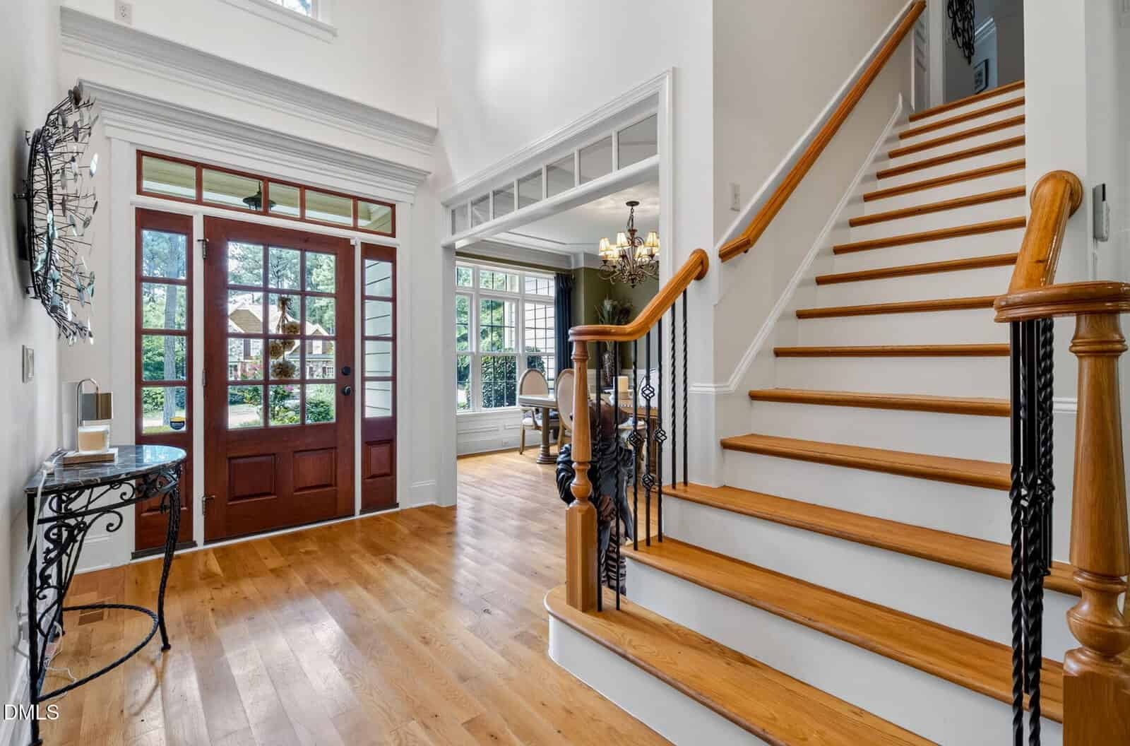A spacious foyer with hardwood floors, a wooden staircase with black railings, and large front doors with glass panels. Natural light fills the area, revealing a bright dining room to the right.