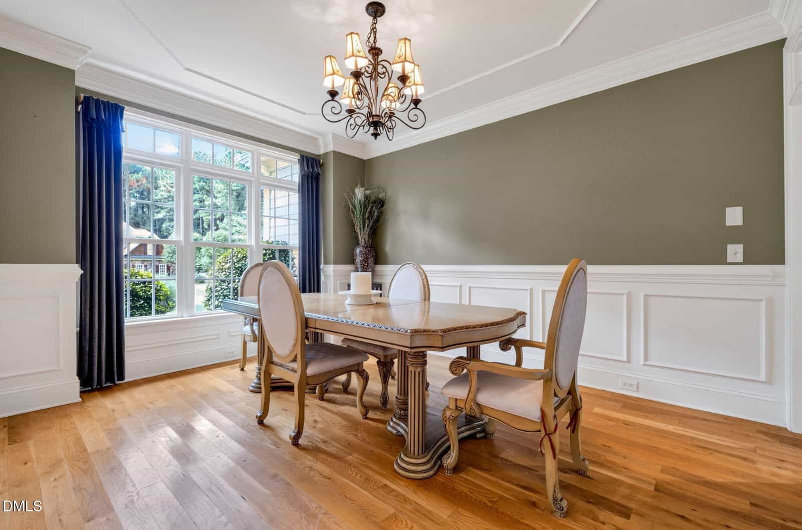 Elegant dining room with wood floors, olive-green walls, white wainscoting, a chandelier, and a large window letting in natural light. Oval table with four chairs and a decorative vase in the corner.