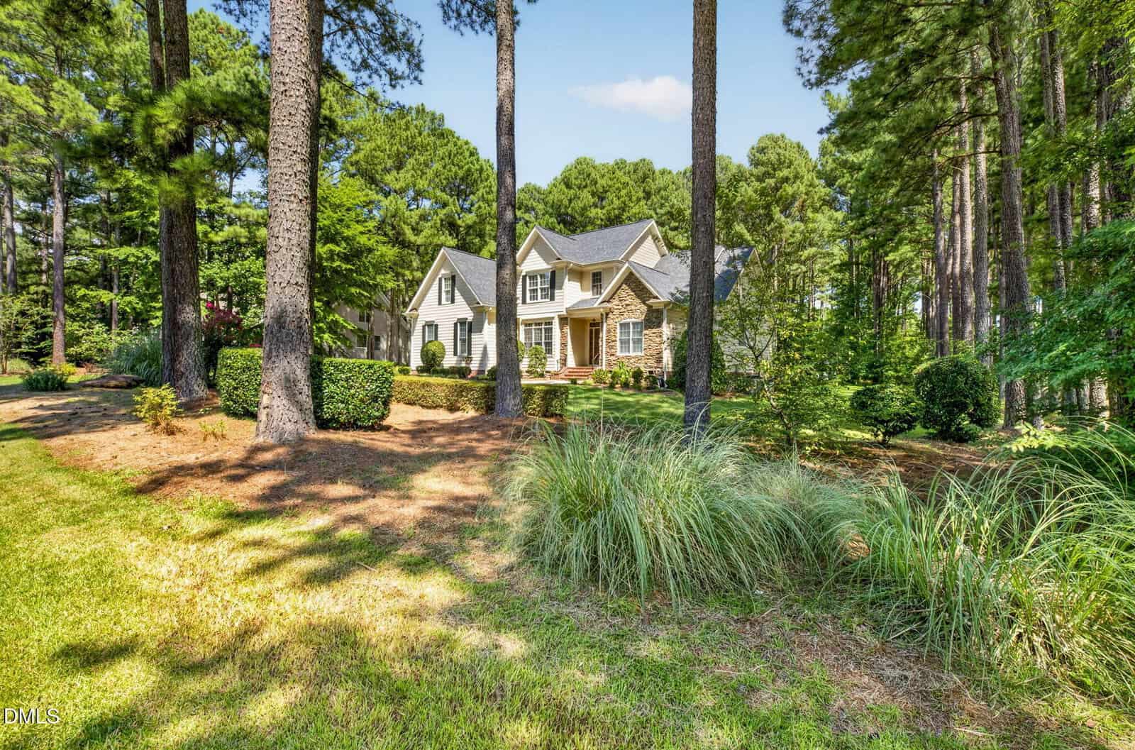 Two-story house with gray siding and white trim is surrounded by tall pine trees, shrubs, and patches of grass on a sunny day. The house features multiple gables and a front porch with columns.