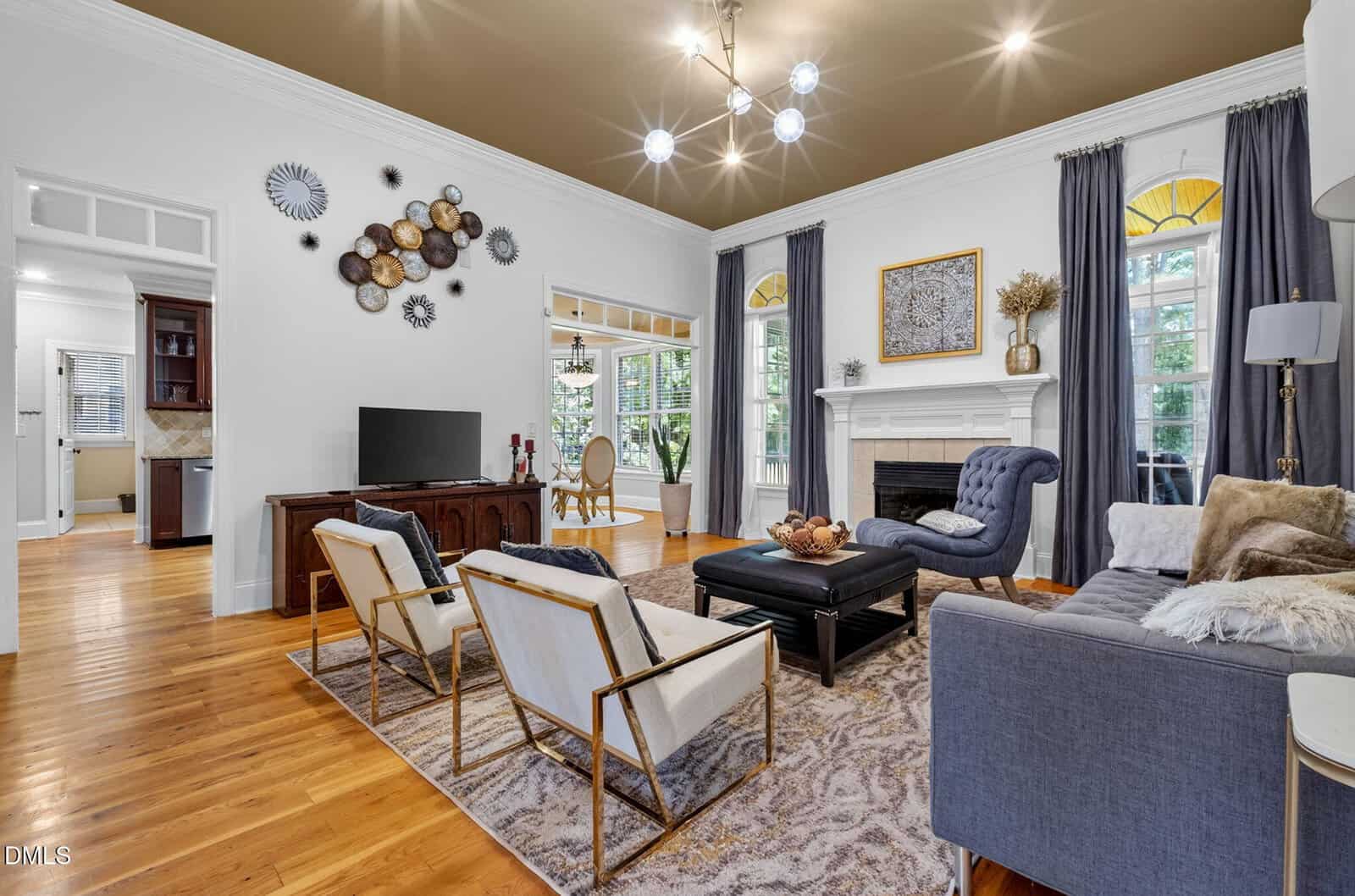 Elegant living room with modern furniture, gold accents, a fireplace, TV console, large windows with blue curtains, hardwood floors, and an open doorway leading to a dining area. Contemporary light fixture overhead.