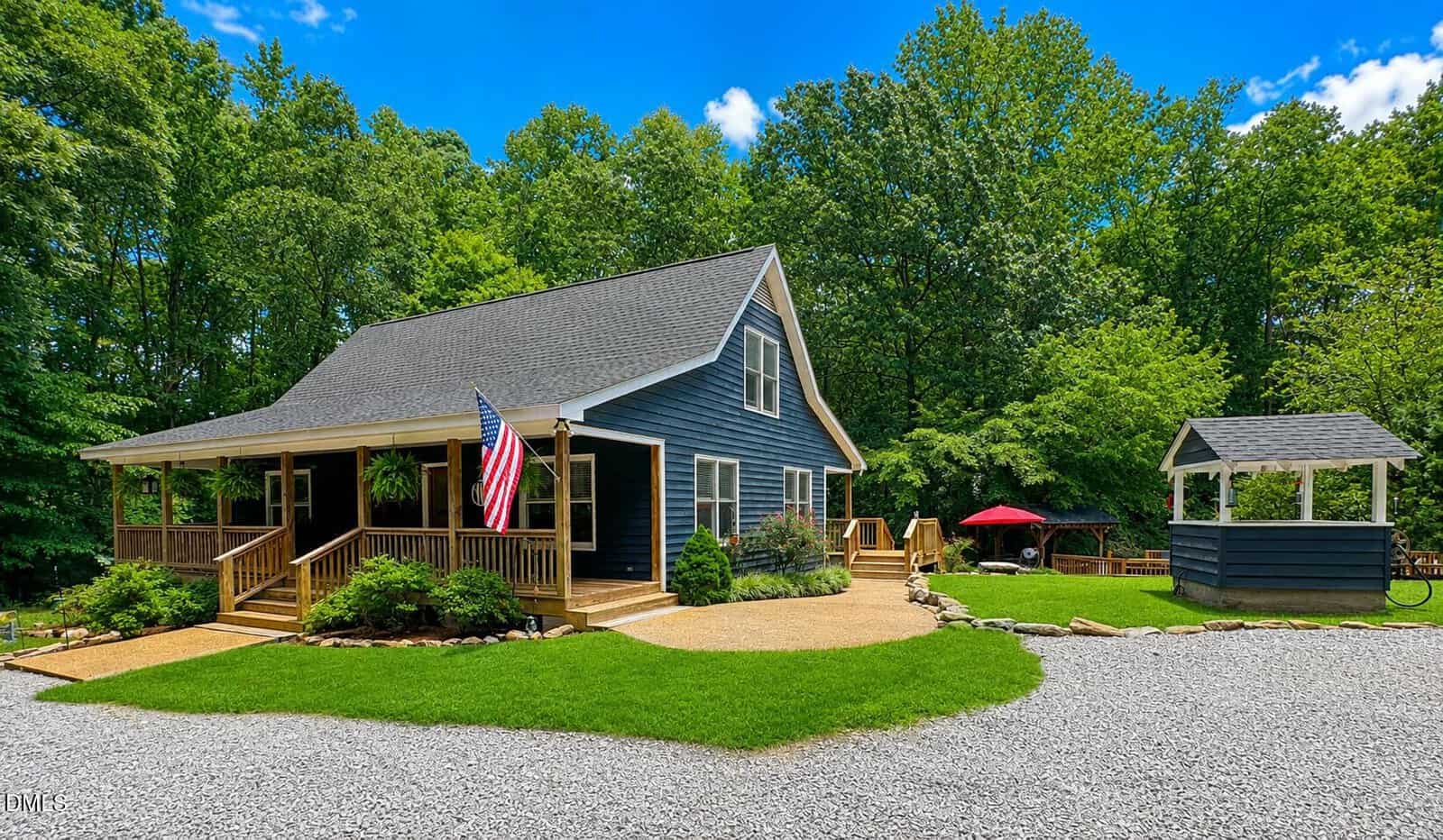 A dark blue house with a front porch and American flag is surrounded by green lawn and trees. A gravel driveway leads to the house. A small open-sided structure and red umbrella are visible in the yard.