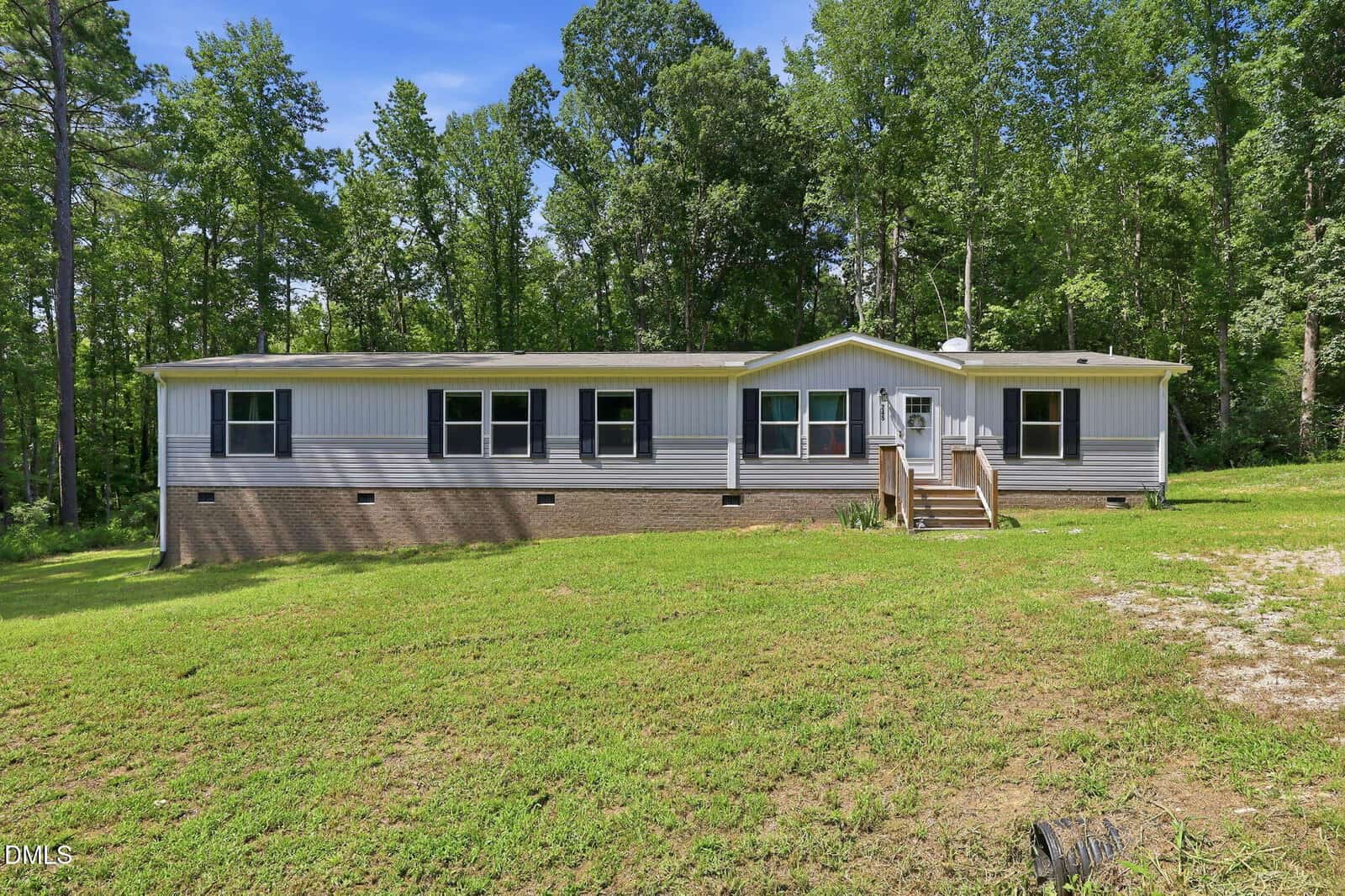 Single-story manufactured home with beige siding and a brick foundation, set on a grassy lawn with scattered trees and a wooded background in Franklinton. A small set of steps leads to the front door at 245 Oak Forest Lane. The sky is clear and sunny.