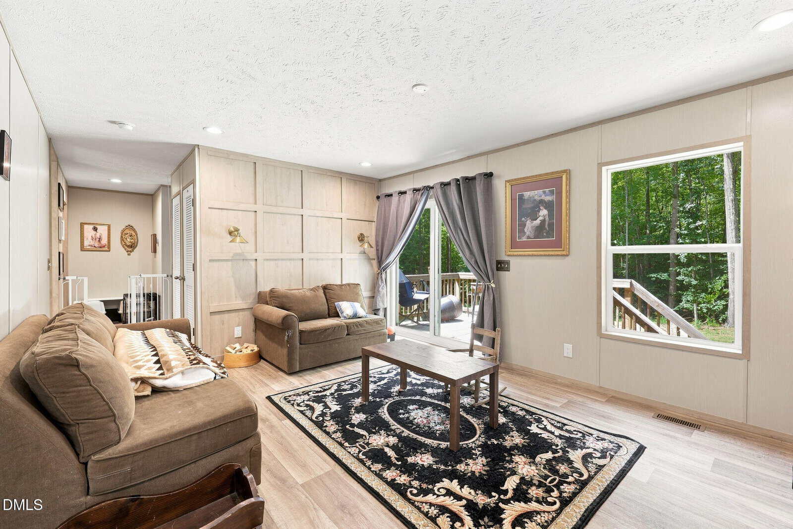 Living room with two brown sofas, a black patterned rug, a wooden coffee table, wall art, and large window with curtains overlooking a deck and green trees. Neutral walls and light wood floors complete the space.