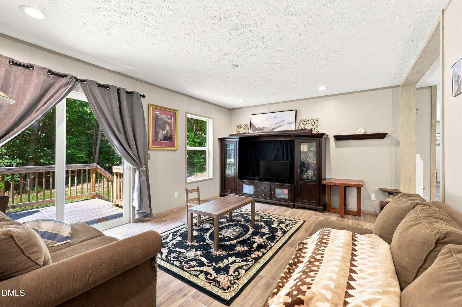 Living room with two couches, a patterned rug, a coffee table, and a dark wood entertainment center with a TV. A sliding door opens to a deck and green outdoor space. Walls are light-colored, and natural light enters the room.
