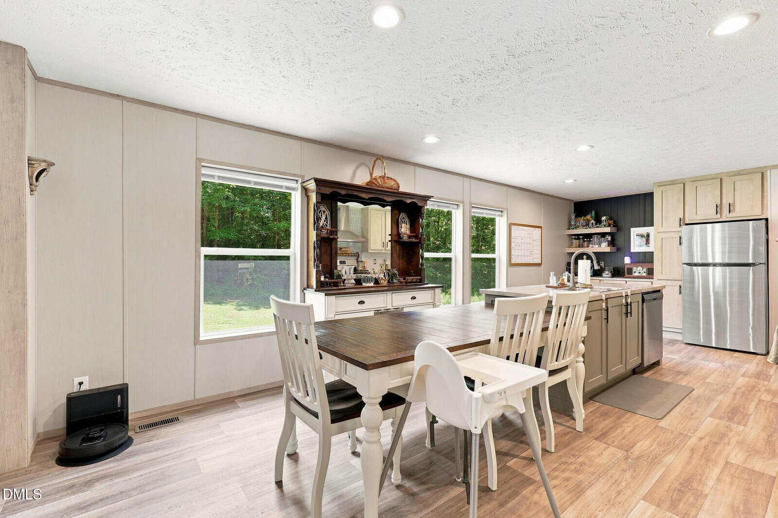 Bright kitchen with wood flooring, white and gray cabinets, a dining table with chairs and a high chair, a hutch against the wall, large windows, and stainless steel refrigerator. A robotic vacuum is near the vent.