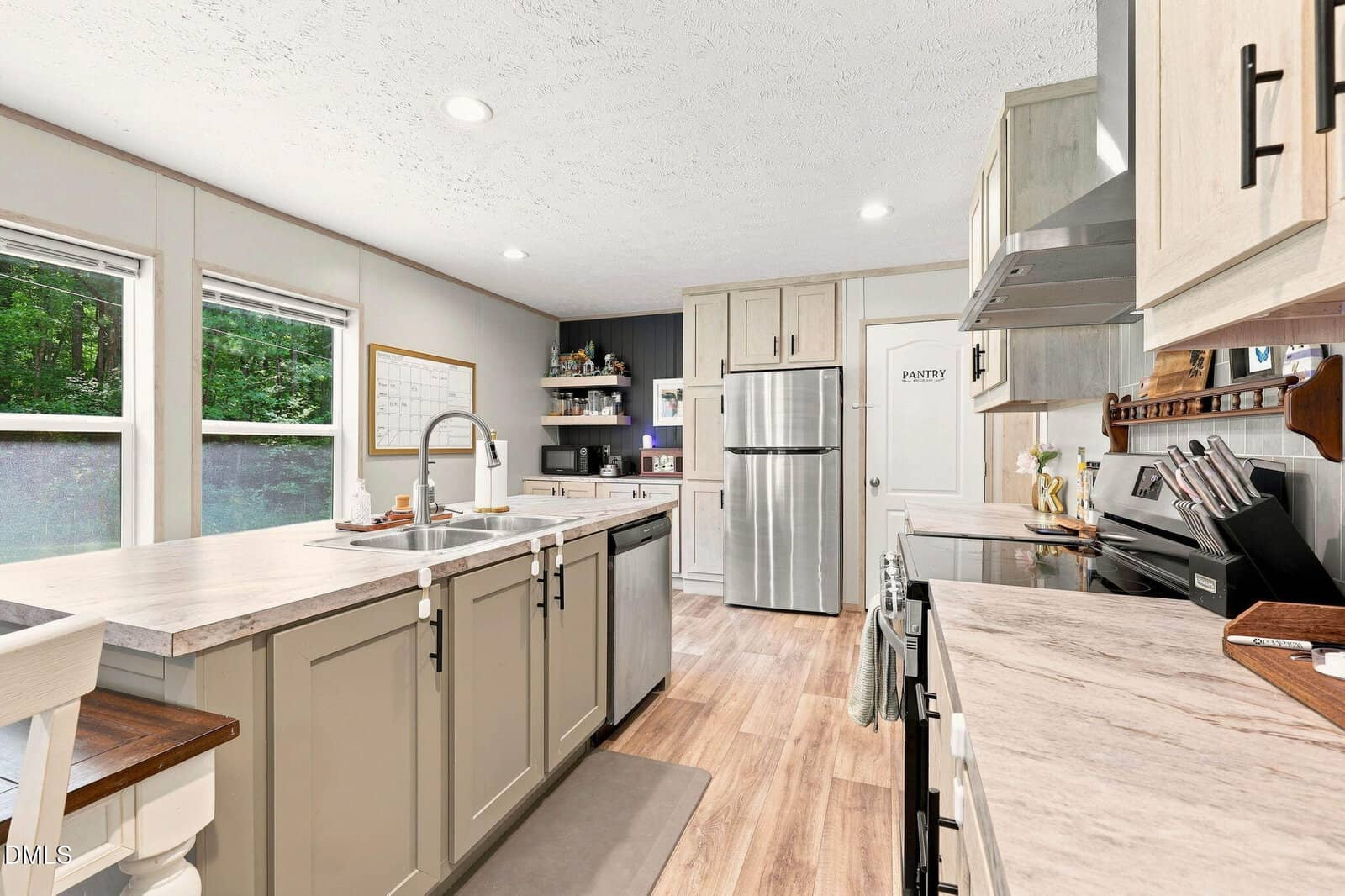 Modern kitchen with neutral cabinets, stainless steel appliances, and wood floors. Large windows let in natural light. A pantry door and open shelves are visible in the background.