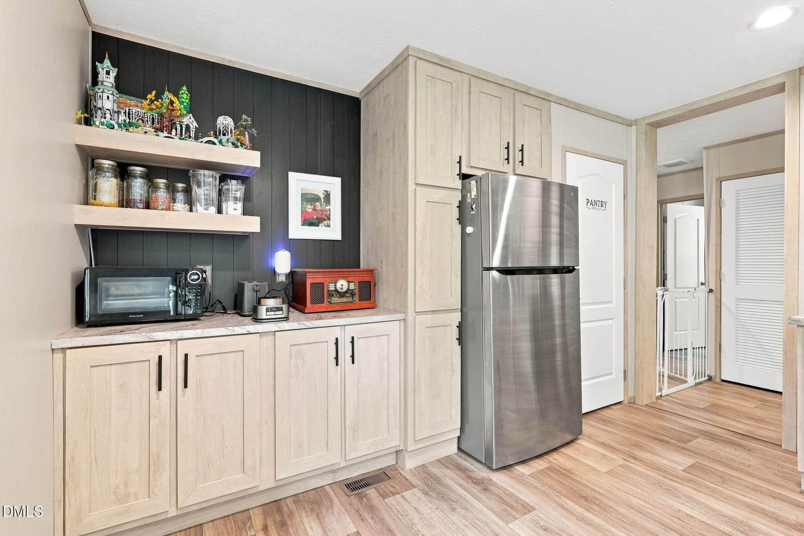 A modern kitchen corner with light wood cabinets, a stainless steel fridge, small appliances, glass jars on shelves, framed art, and wood-look flooring. A door labeled “Pantry” is visible in the background.